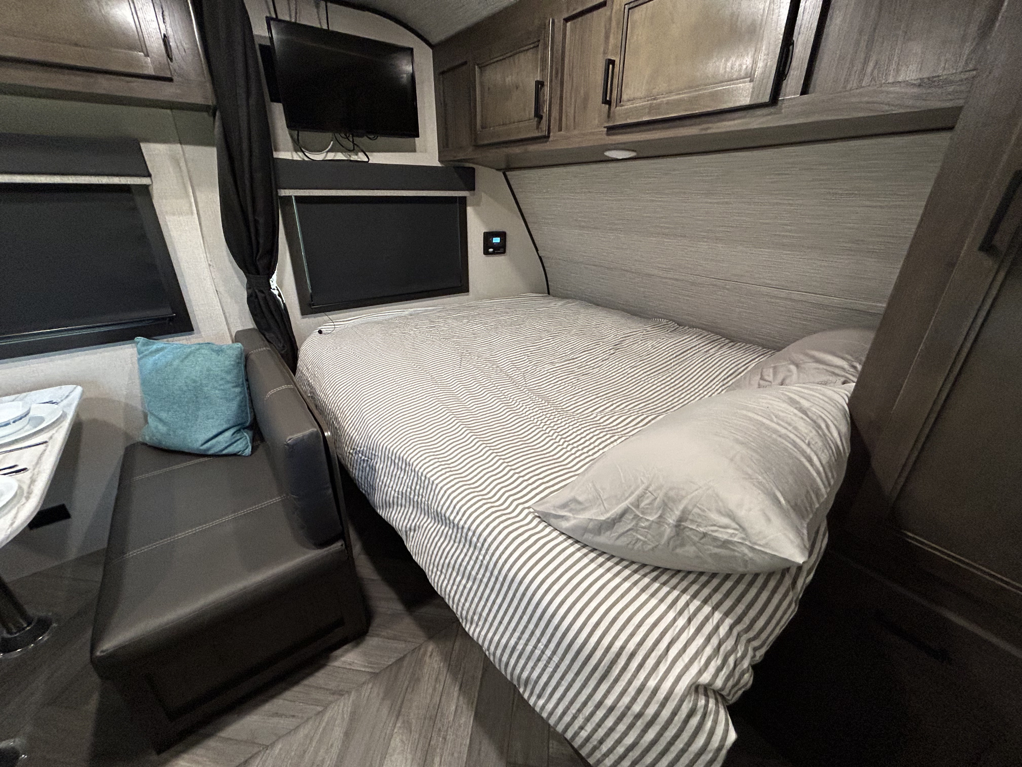 Inside an RV rental, a compact bedroom features a striped bed, overhead cabinets, and a small dinette.. Olympia Sport 2022