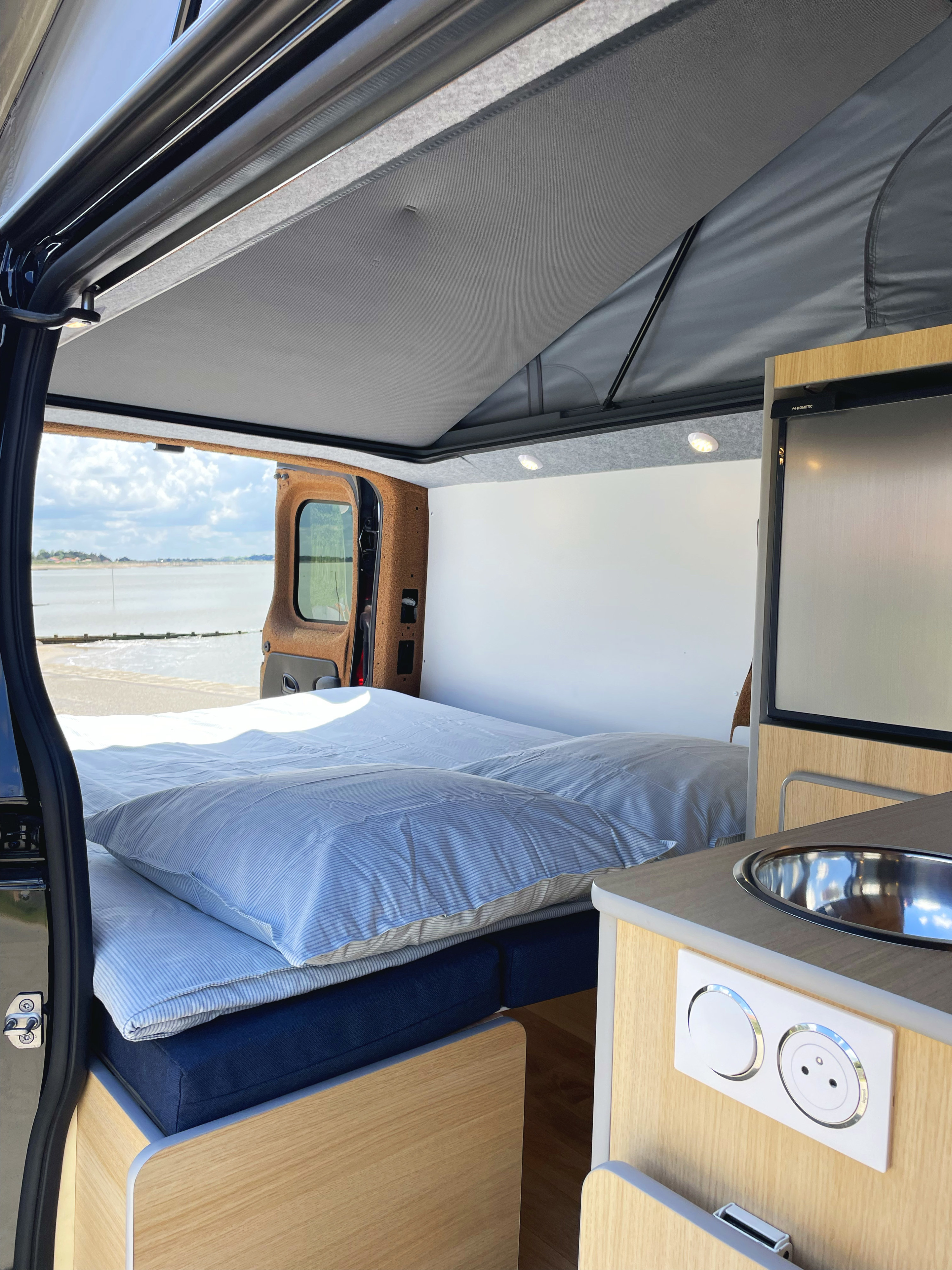 Compact RV rental interior featuring a cozy bed, pop-top roof, and kitchen with sink & fridge. Ready for adventure by the water.. Renault TRAFIC 2022