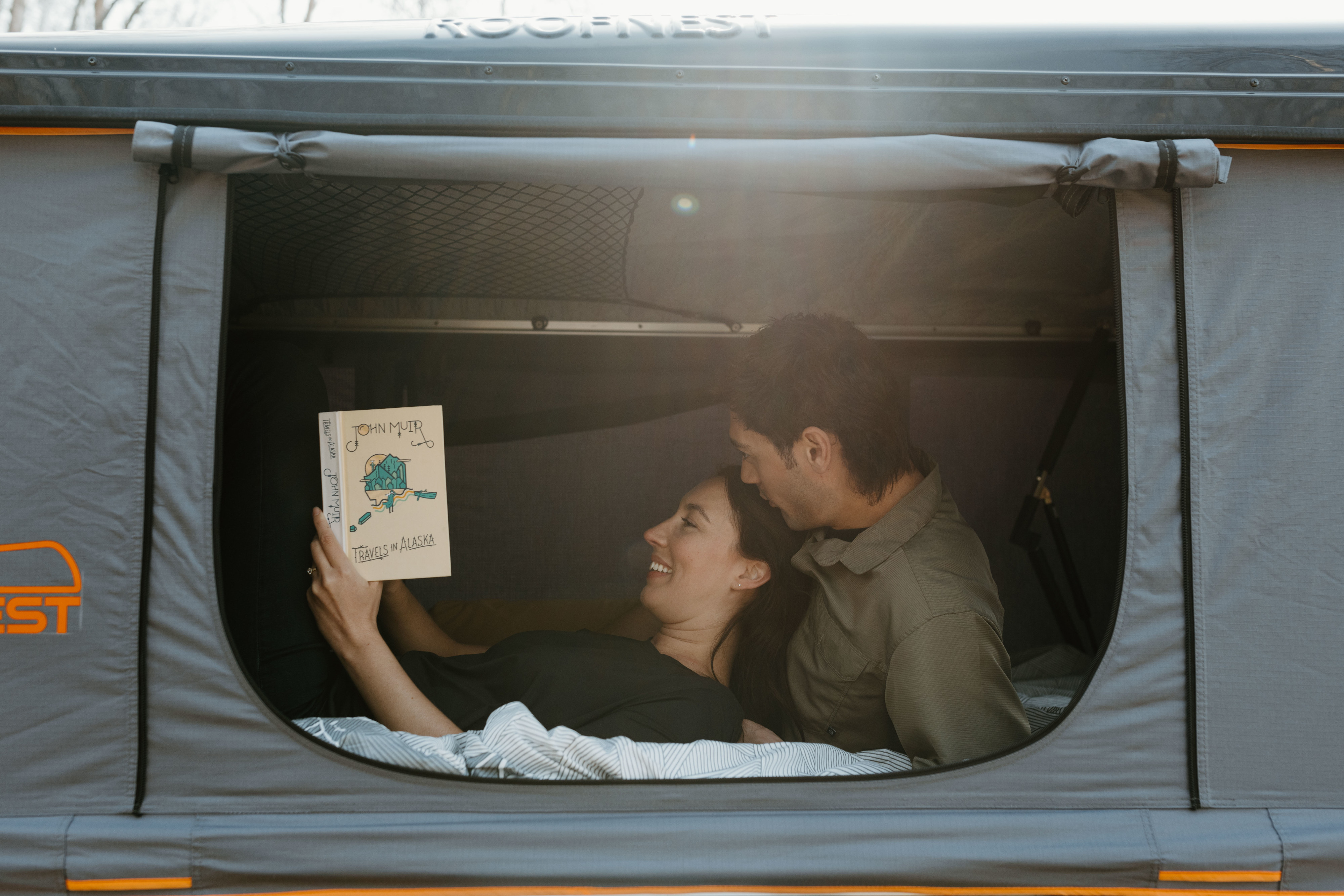 A happy couple relaxes in a Roofnest rooftop tent, reading "Travels in Alaska," ideal for any RV rental adventure.. Jeep Wrangler 2023
