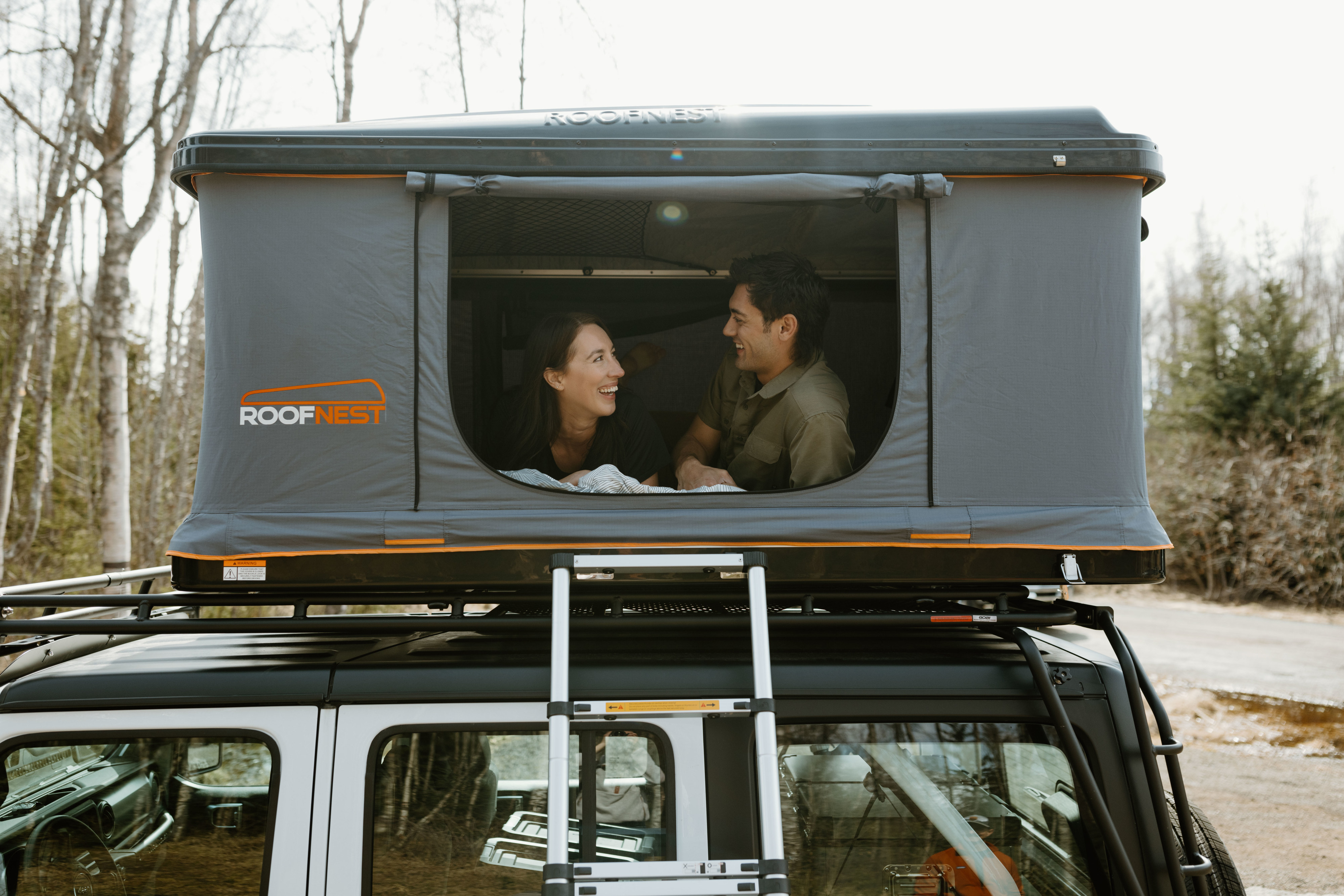 Happy couple in a Roofnest roof-top tent on a white SUV, a unique RV rental option for camping adventures.. Jeep Wrangler 2023
