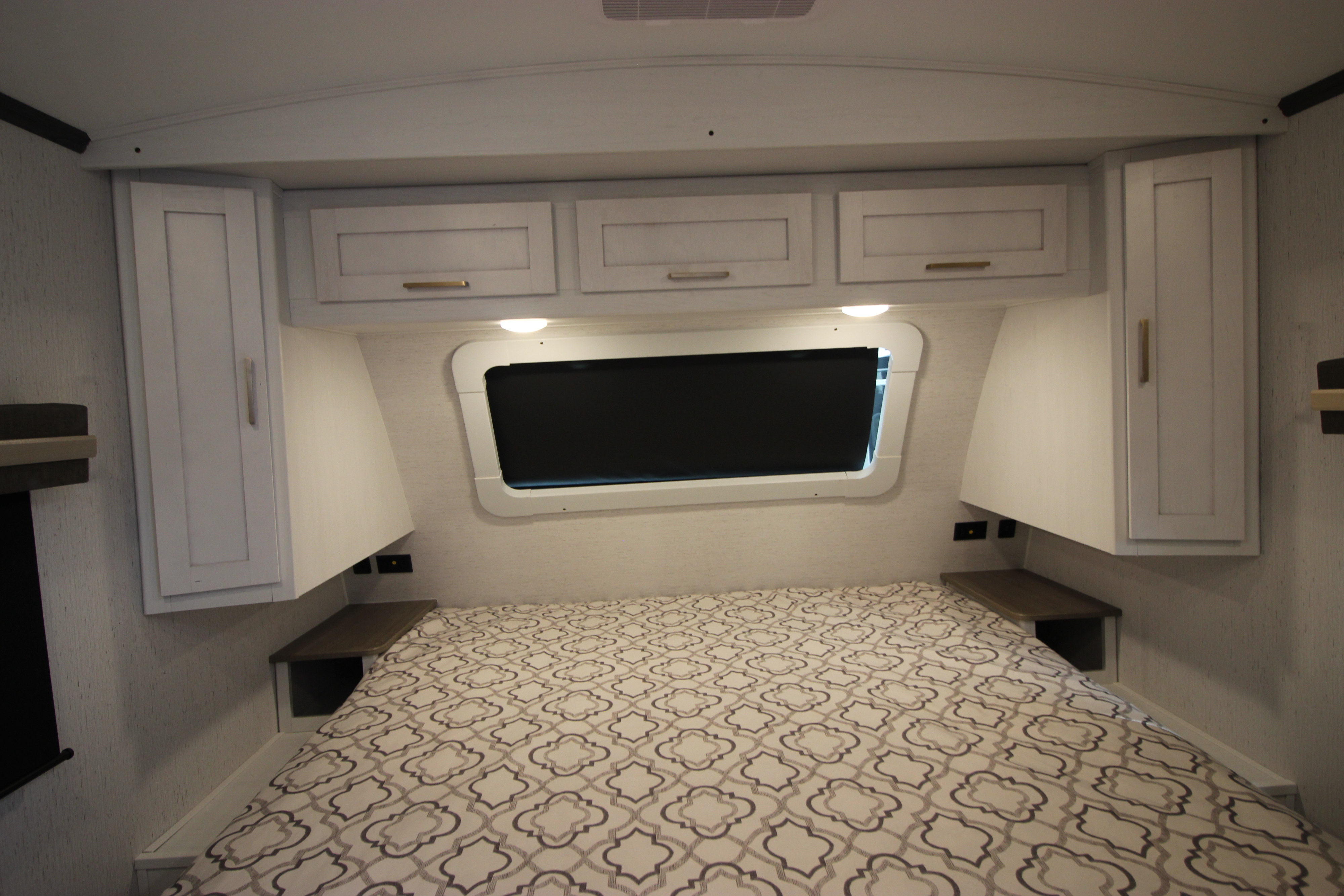 An RV rental bedroom with white cabinets, side shelves, a window, and a patterned bed, ready for adventures.. Dutchmen Astoria 2023