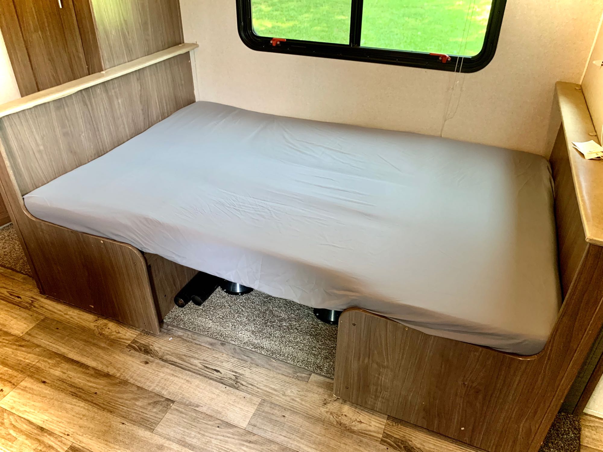 This cozy RV rental interior features a bed with a grey sheet, wood-look paneling, and a green outdoor view through the window.. Keystone RV Hideout LHS 2019