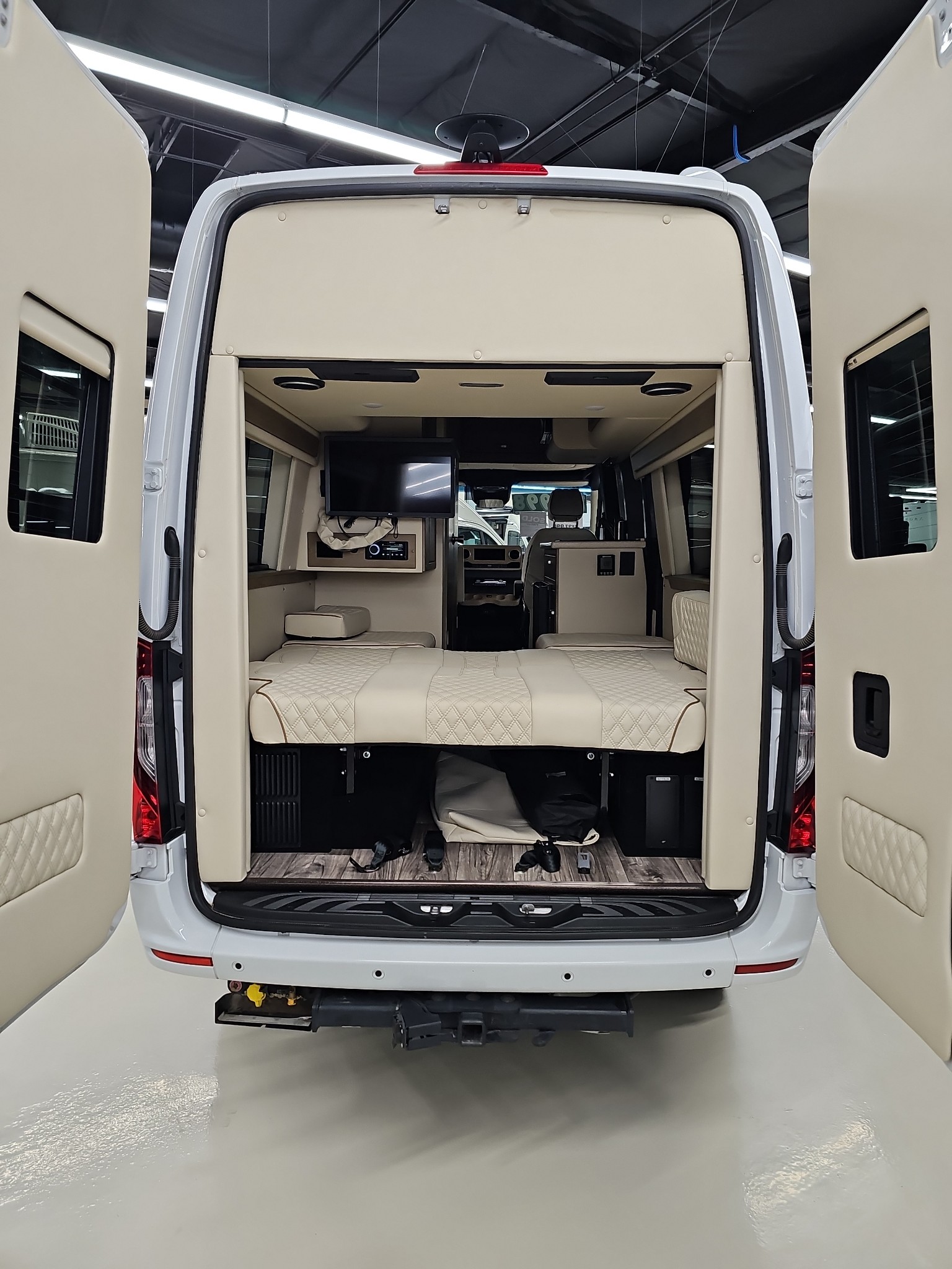 White RV rental from the rear, showing a luxurious interior with open doors, featuring a cream quilted bed, TV, and wood-look floor.. Midwest Automotive Designs Midwest Automotive Designs Class B 2022