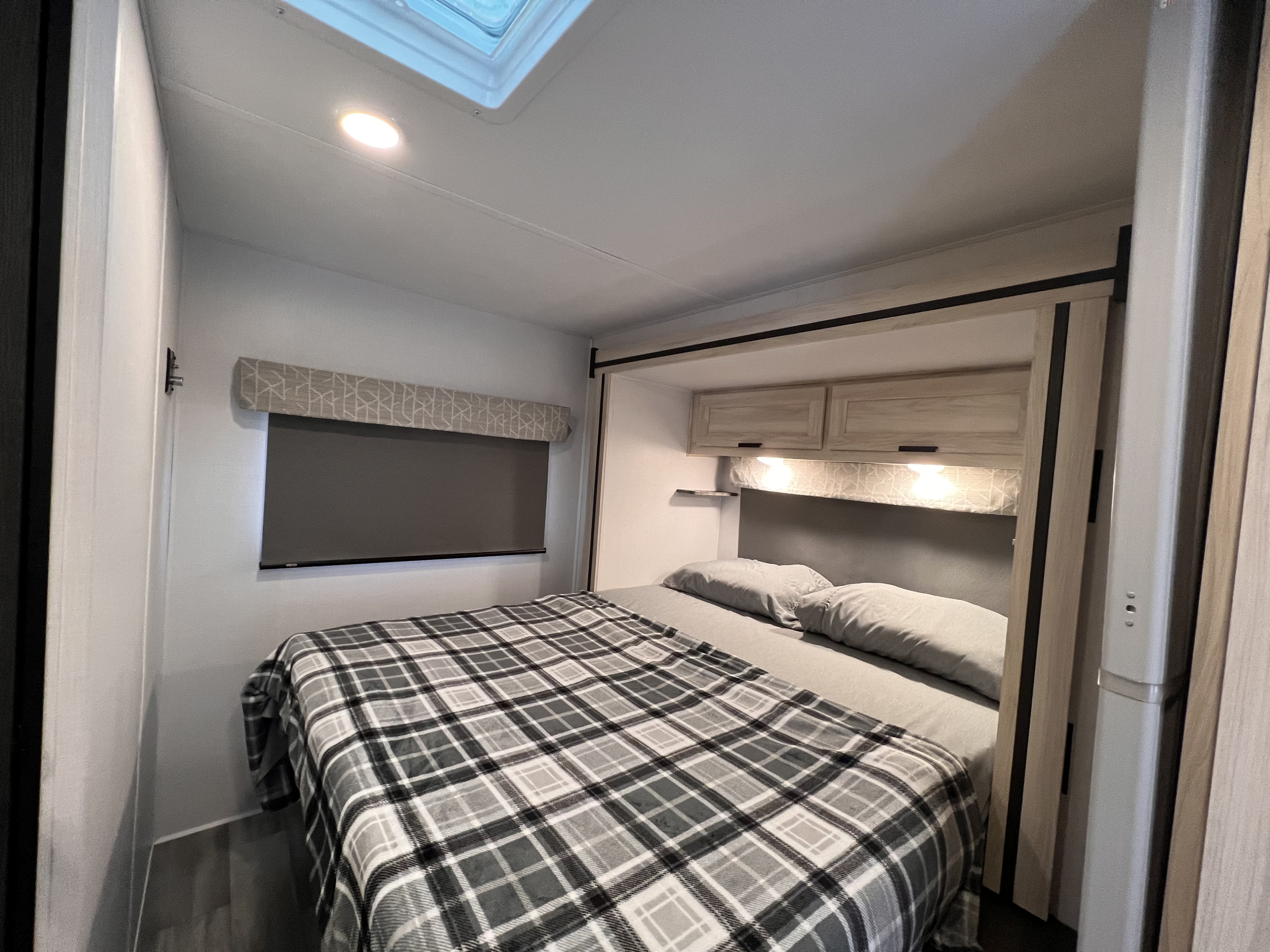 Cozy RV rental bedroom featuring a full bed with plaid blanket, overhead storage, skylight, and window shade.. Forest River Sunseeker 2024
