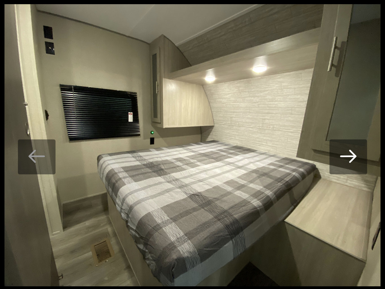 This RV rental bedroom features a comfy bed with grey plaid bedding, overhead cabinets, and a window with blinds.. Coachmen Catalina 2023