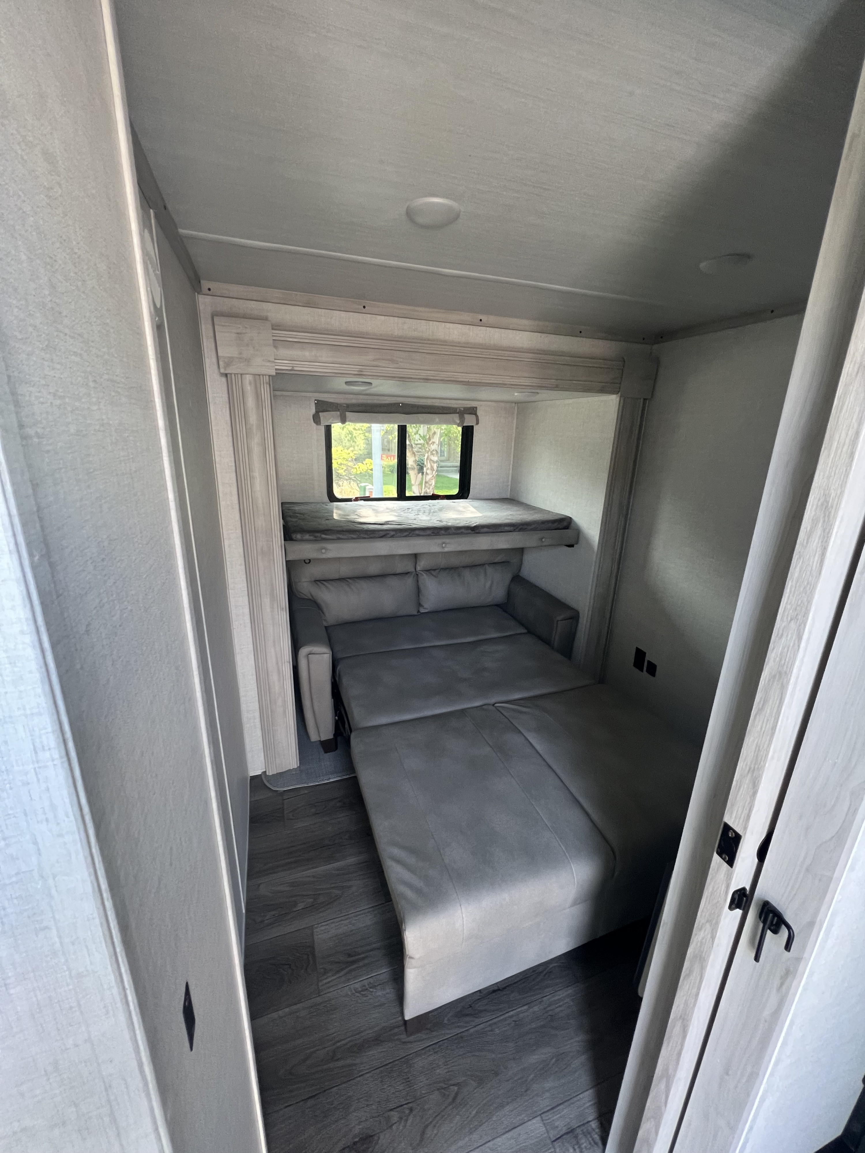 Modern RV rental interior with a gray convertible sofa, upper bunk, and window.. East to West Tandara 2022