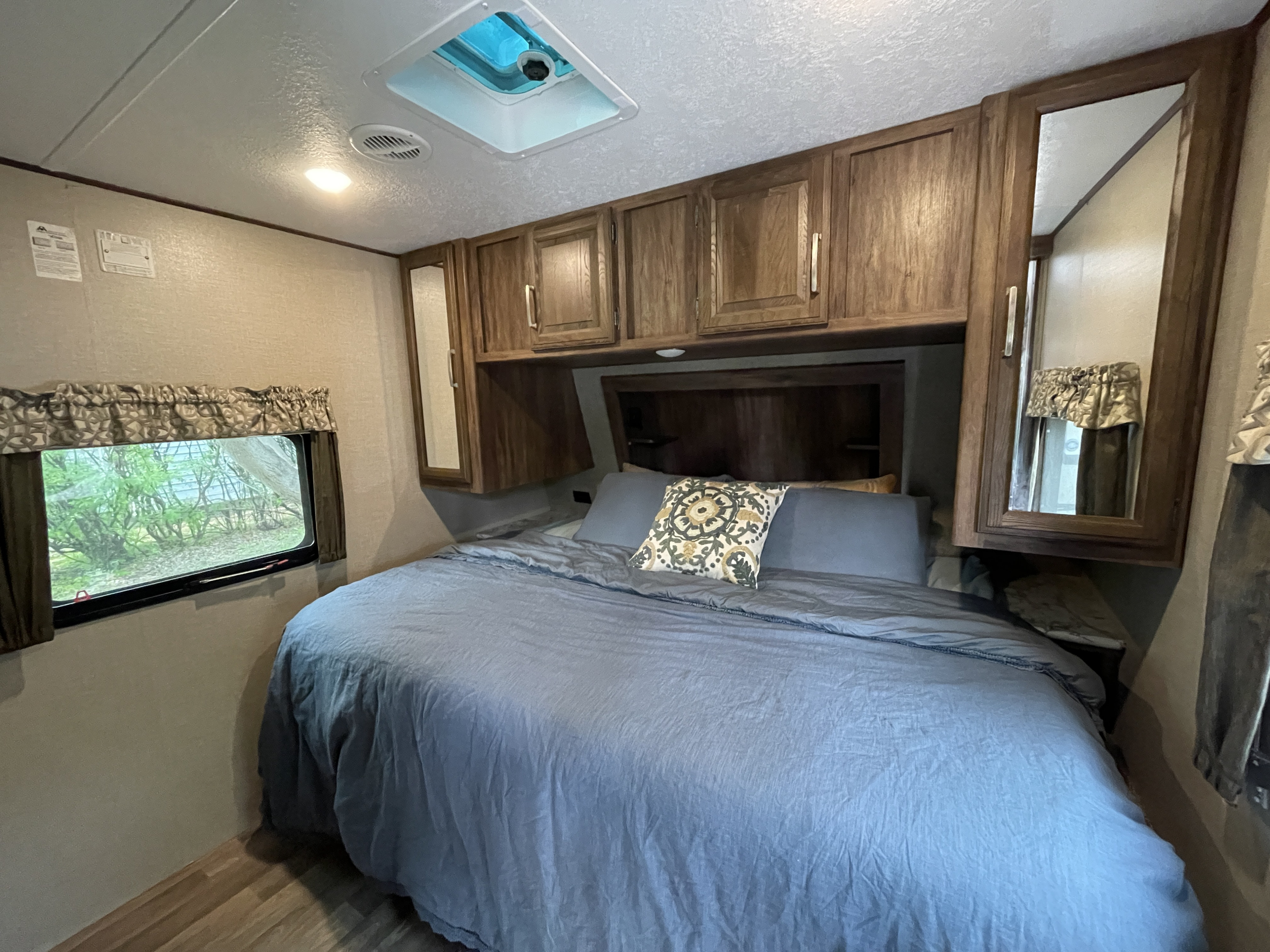RV rental bedroom: a cozy space with a large bed, overhead wood cabinets, and a window offering a lush green view.. Keystone RV Cougar 2018