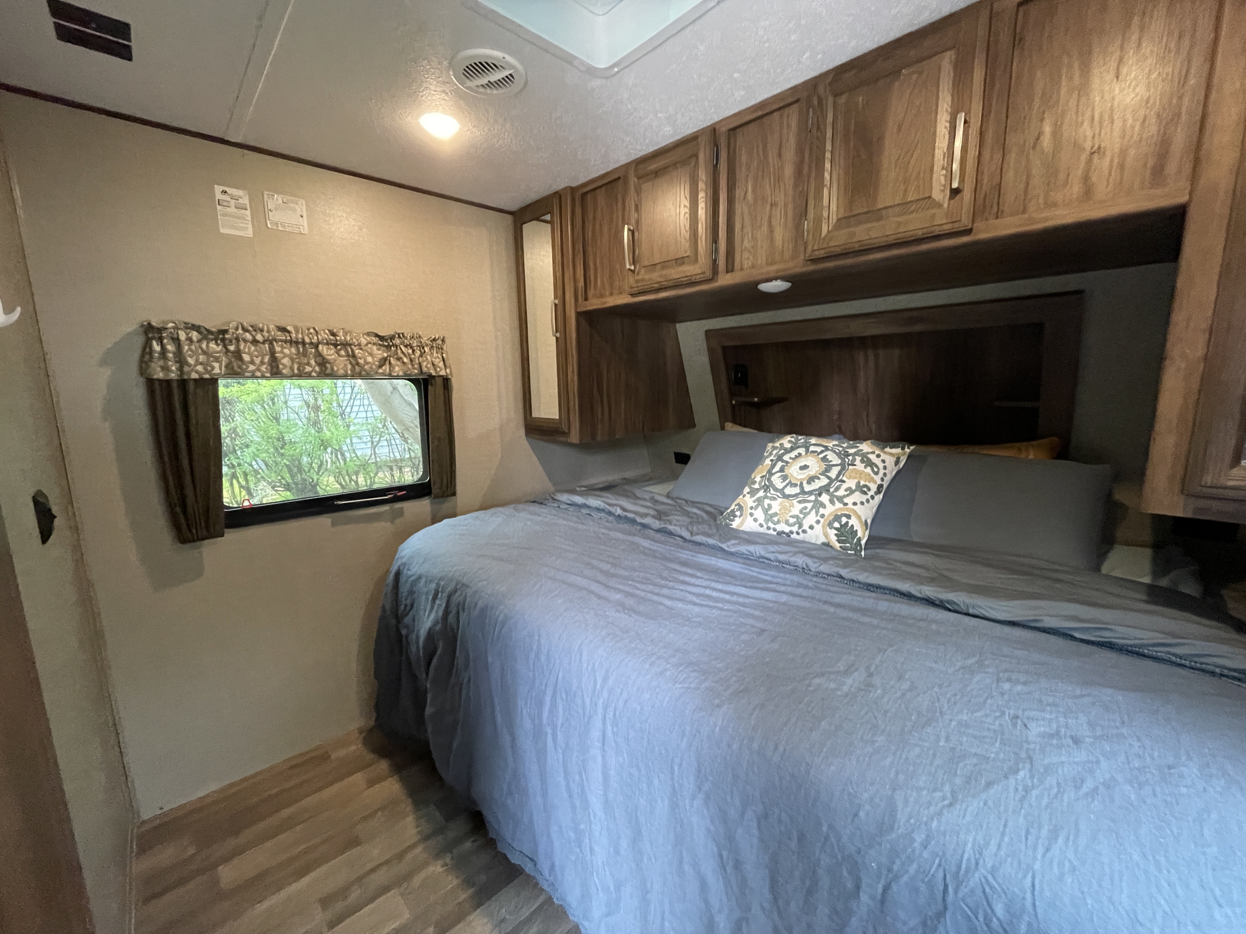 Cozy RV rental bedroom with light blue bedding, a decorative pillow, overhead wood cabinets, and a window overlooking trees.. Keystone RV Cougar 2018