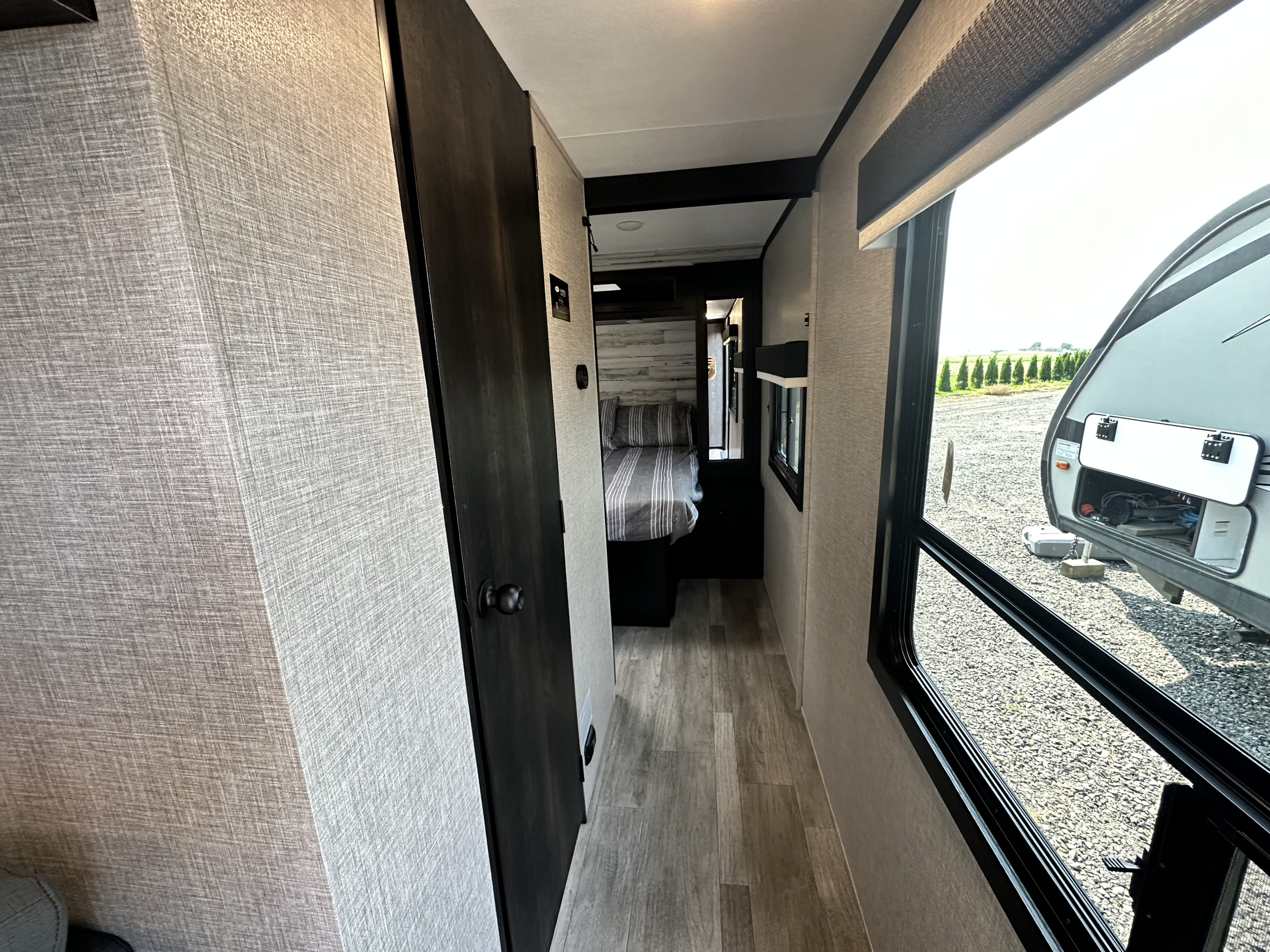 View inside an RV rental showing a hallway, bedroom, and window with a view of a gravel lot and exterior storage.. Jayco 30QB 2023