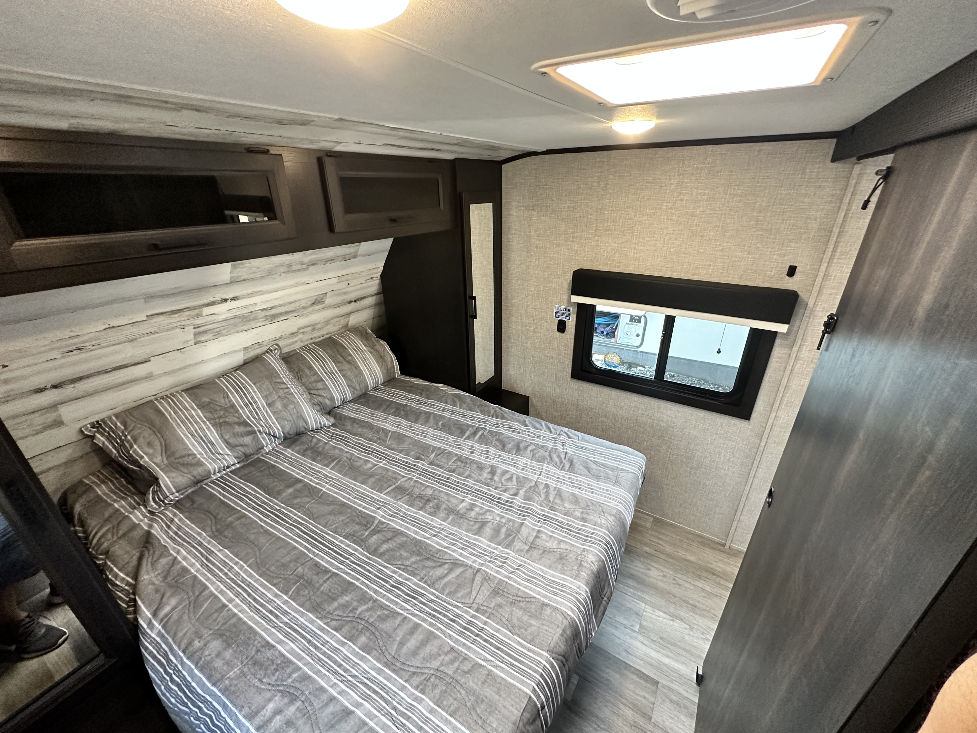 Clean, modern RV rental bedroom with a comfortable striped bed, overhead storage, window, and a mirrored closet.. Jayco 30QB 2023