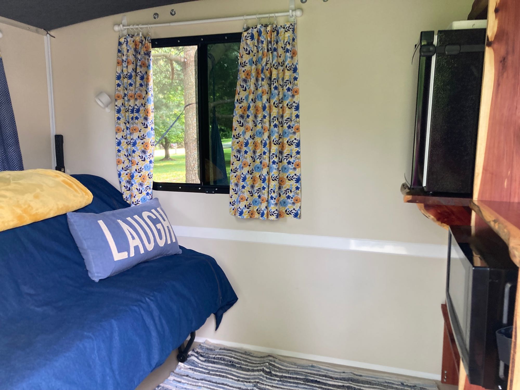 Inside an RV rental: a comfy bed, bright window with floral curtains, & kitchenette.. Runaway Rouser 2023