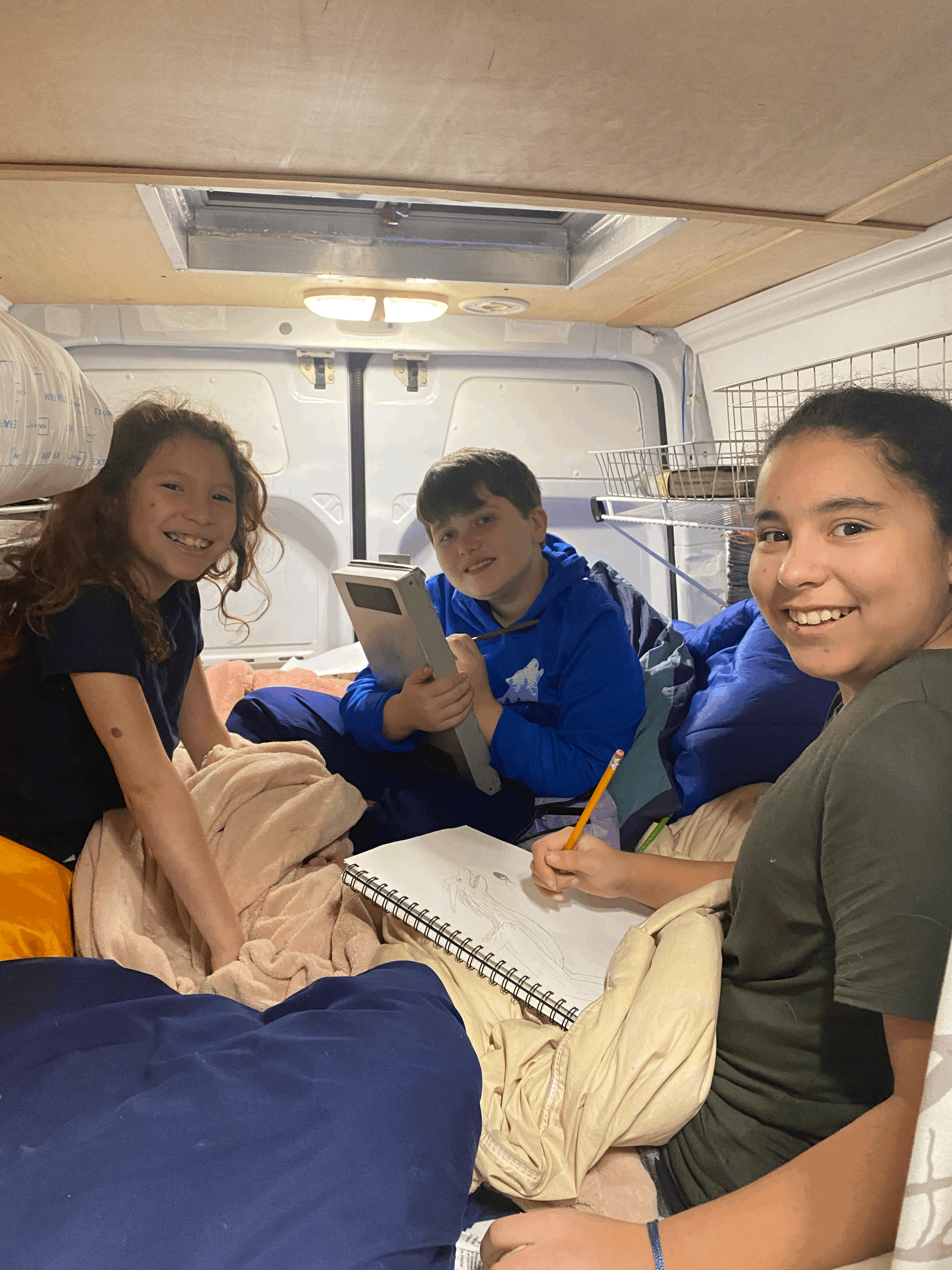 Three smiling kids cozy up in an RV rental, one drawing, others relaxing in the camper van interior.. Mercedes-Benz Sprinter 2017