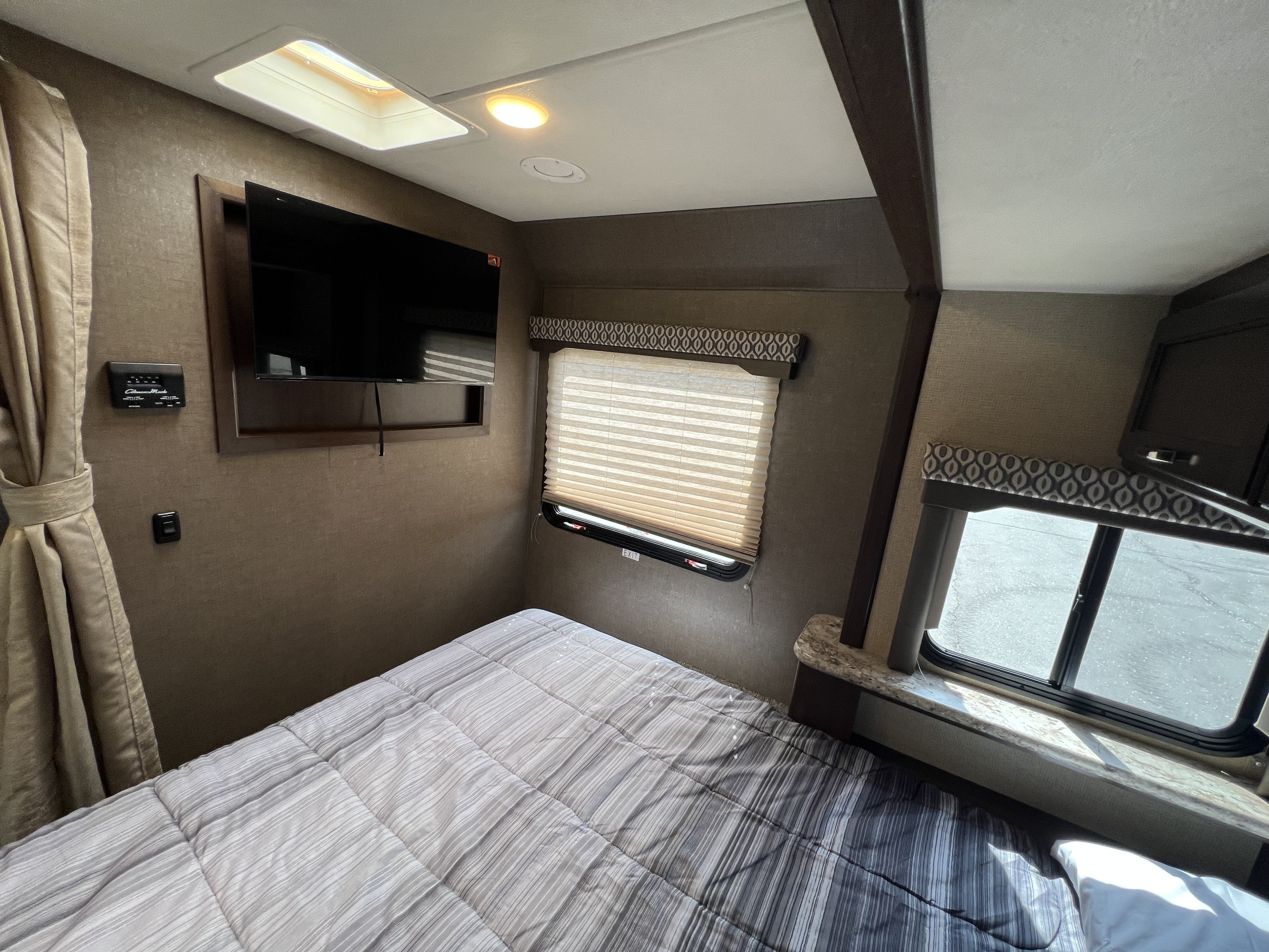 An RV rental bedroom features a bed with striped comforter, wall-mounted TV, window with blinds, and ceiling light.. Thor Motor Coach Freedom Elite 2018