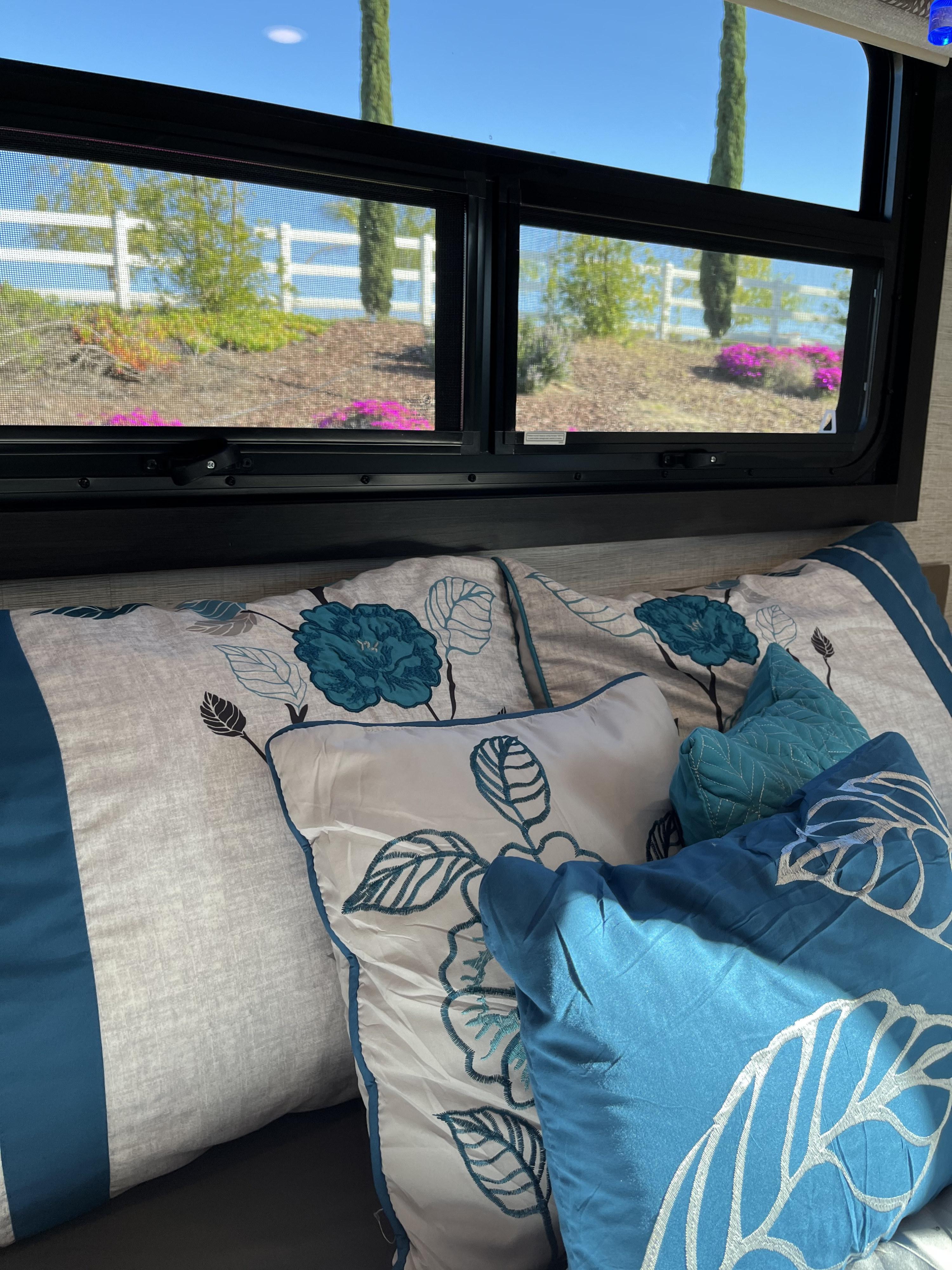 Enjoy the comfort of an RV rental bedroom with a peaceful window view of a white fence, green trees, and pink flowers on a sunny day.. Entegra Coach Esteem 31F 2021