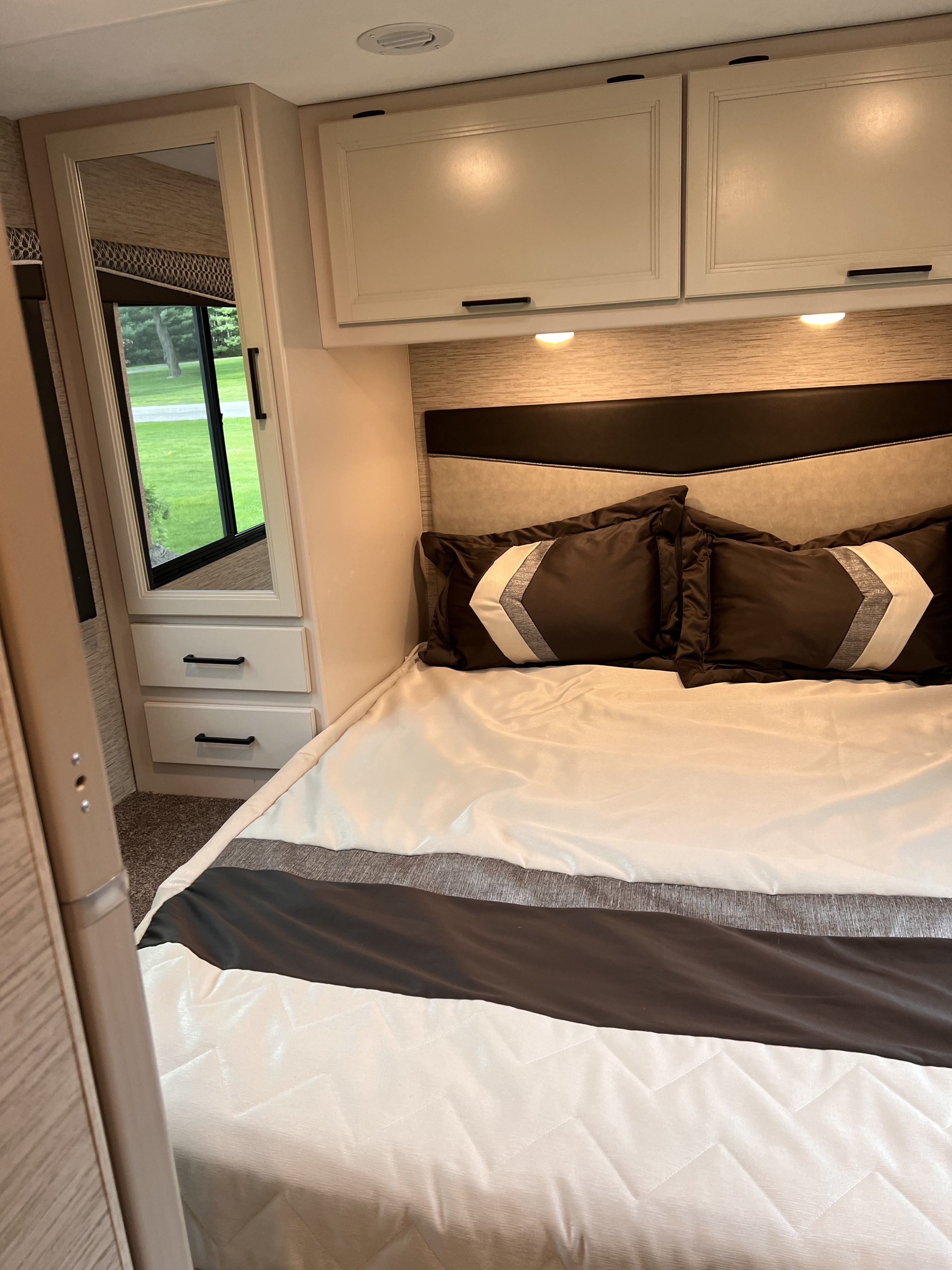 RV rental's inviting bedroom: neutral and dark bedding, overhead cabinets, and a window revealing greenery.. Thor Four Winds 2023