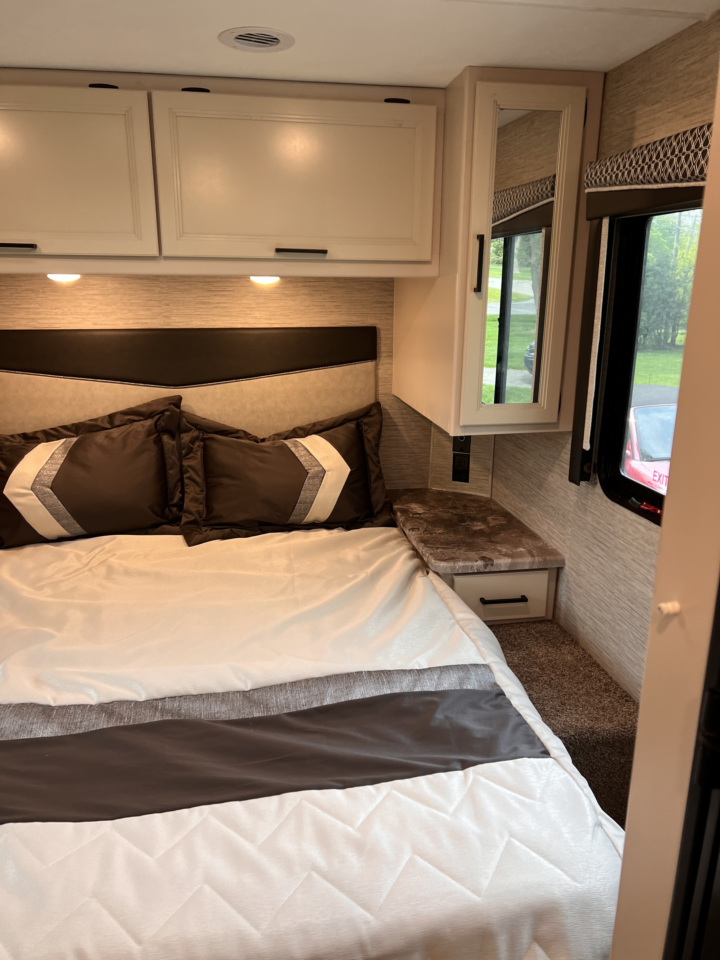 Cozy RV rental bedroom with beige overhead cabinets, a comfortable bed, and a side nightstand with a window.. Thor Four Winds 2023