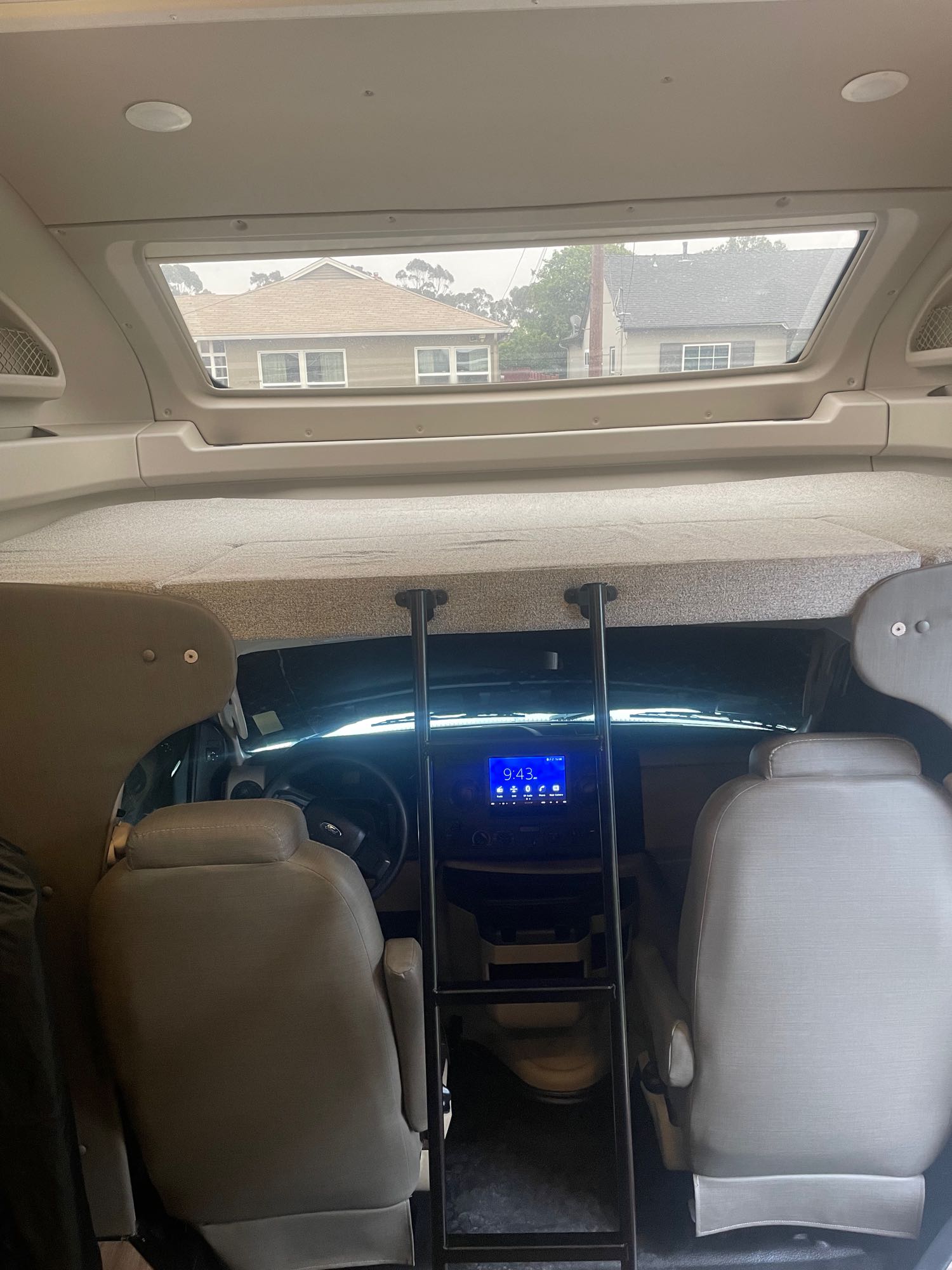 Inside an RV rental, showing the front cab with dashboard, overhead bunk with a ladder, and houses visible through the windshield.. Entegra Coach Odyssey 2021