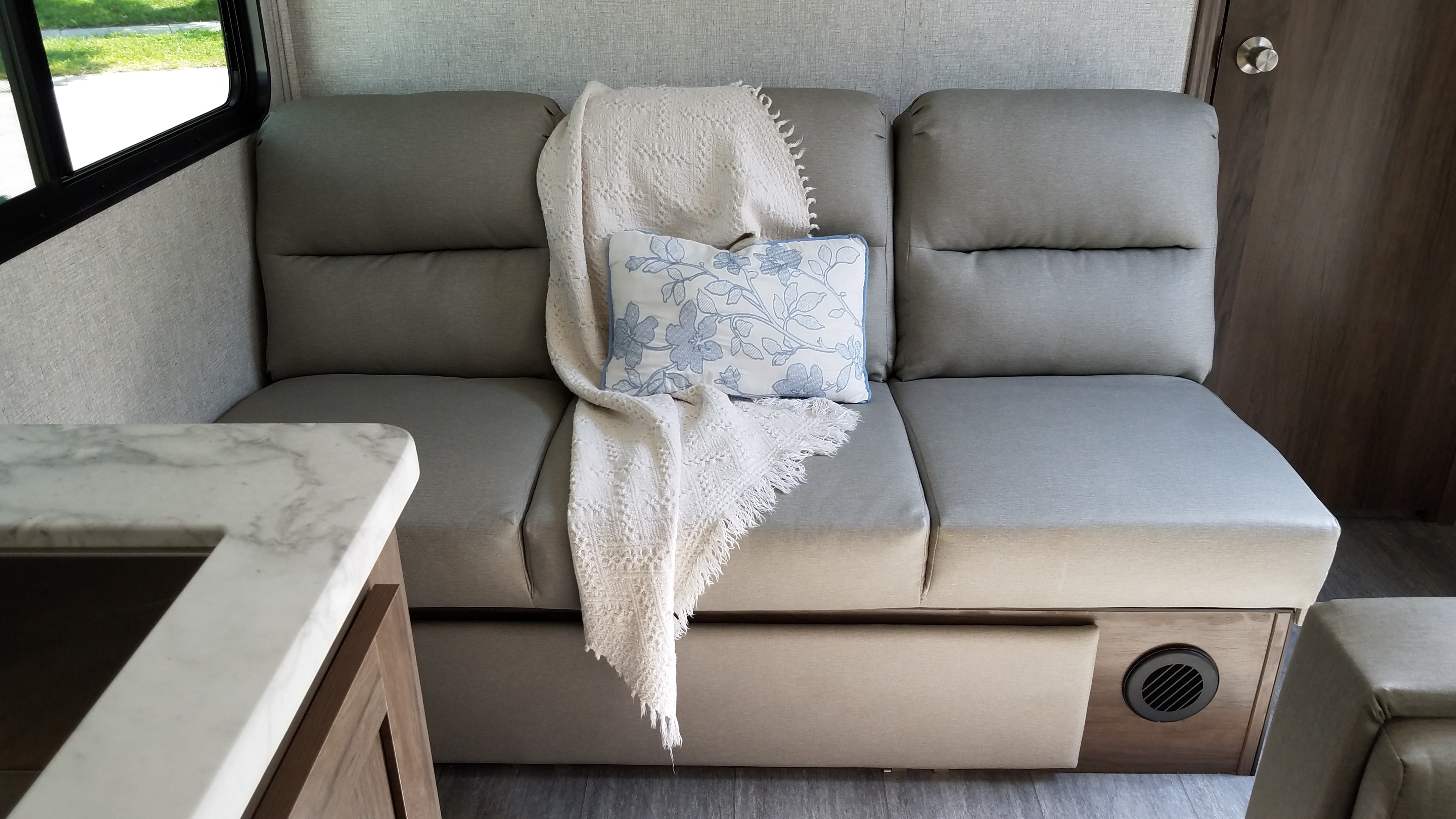 Cozy RV rental living area: a grey sofa with a white textured throw and blue floral pillow, next to a window and marble counter.. East to West Silver Lake 2021
