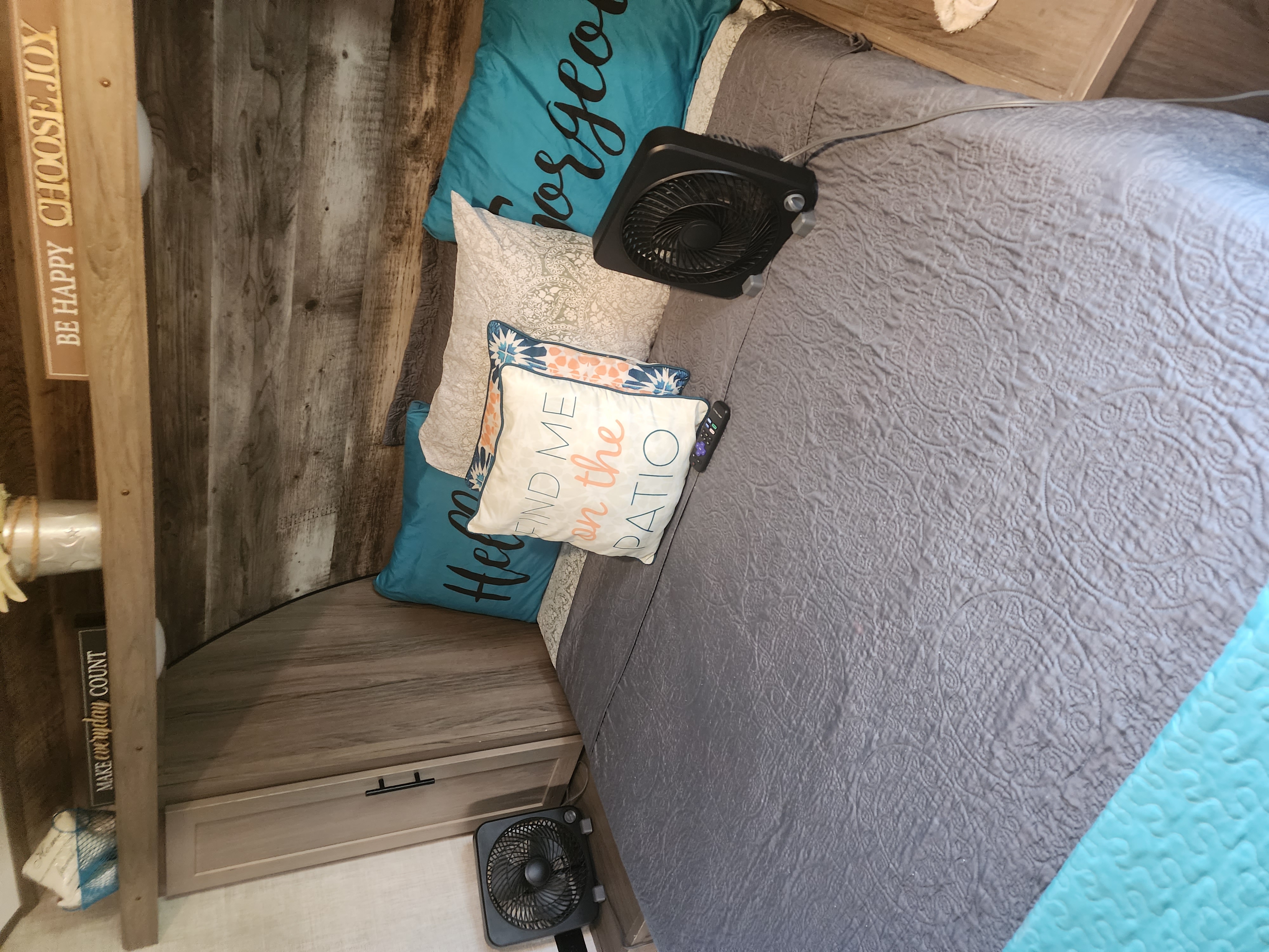 RV rental bedroom featuring a bed with gray quilted bedding, decorative pillows (one says "FIND ME on the PATIO"), a small fan, and wood walls.. Gulf Stream Kingsport 2022