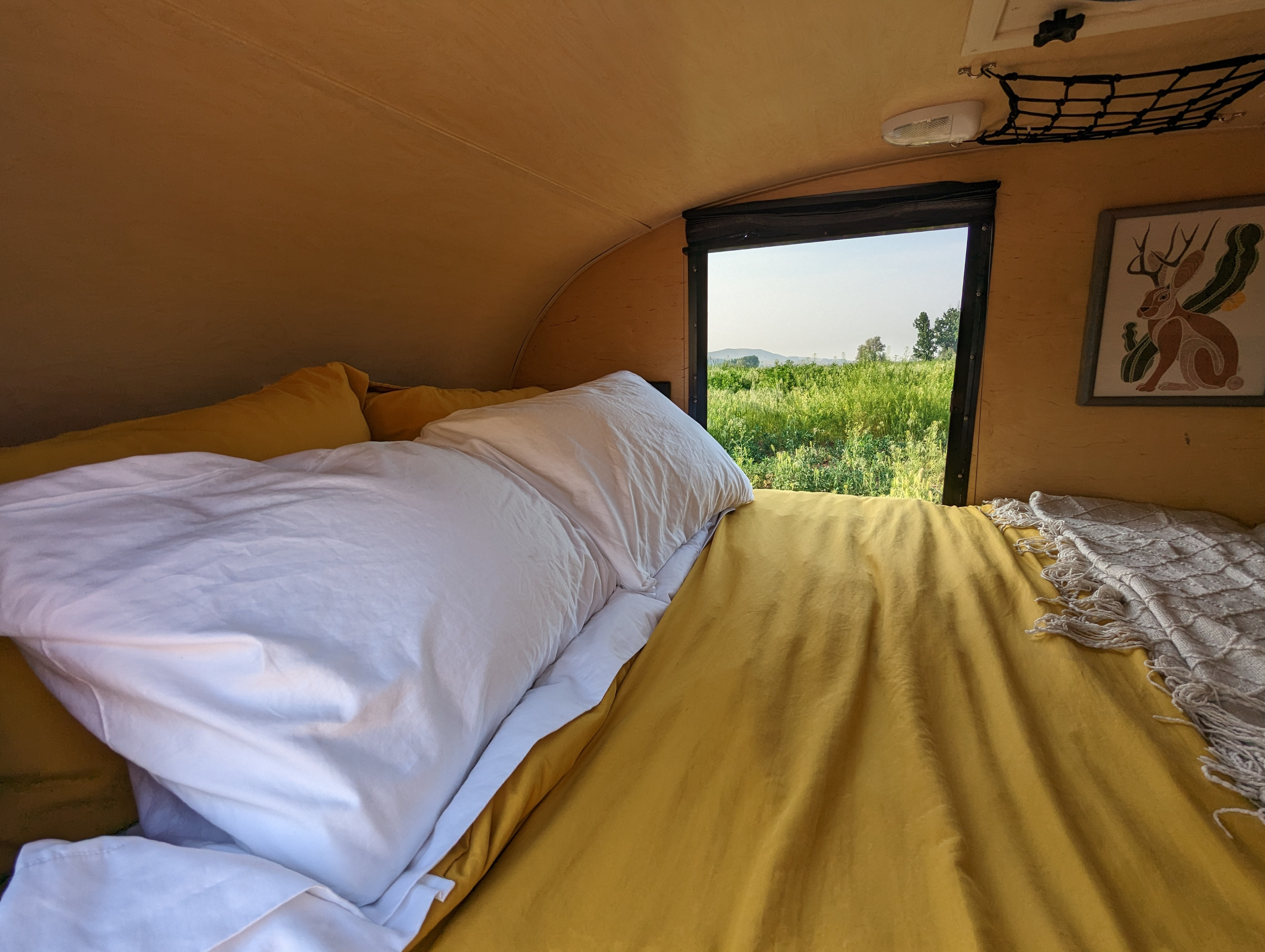 Inside a cozy RV rental: a bed with yellow sheets and white bedding, with a window overlooking a green field.. Little Guy Silver Shadow 2011