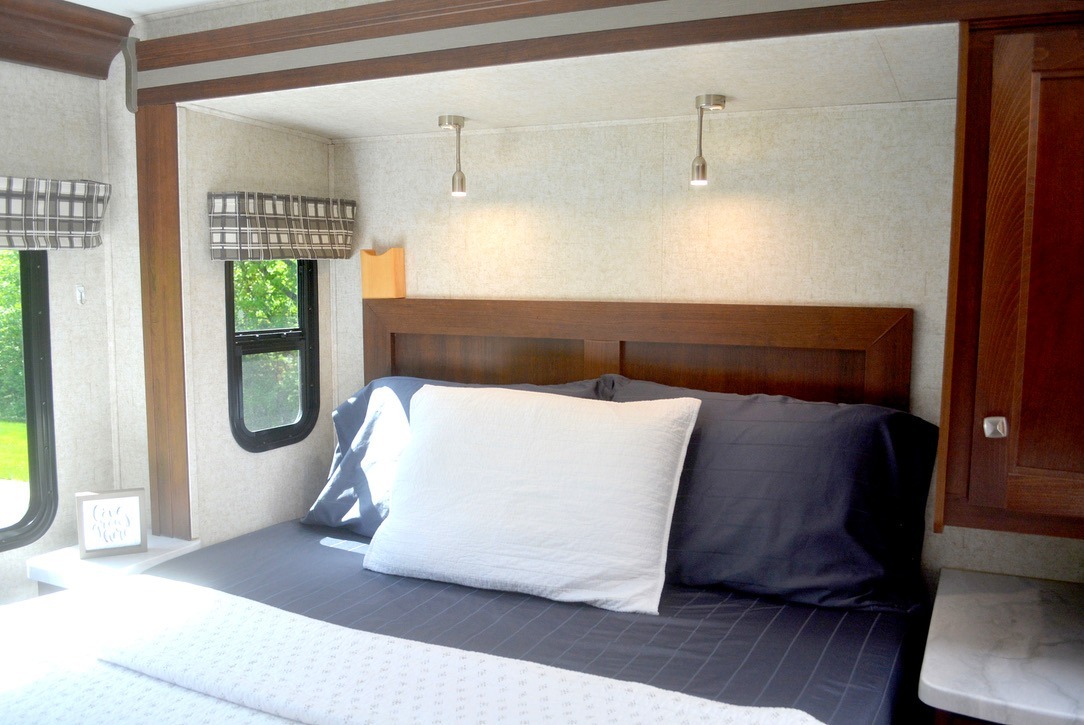 This RV rental bedroom features a comfy bed with navy sheets, a wood headboard, and two small windows with plaid shades.. Forest River Sunseeker 2019