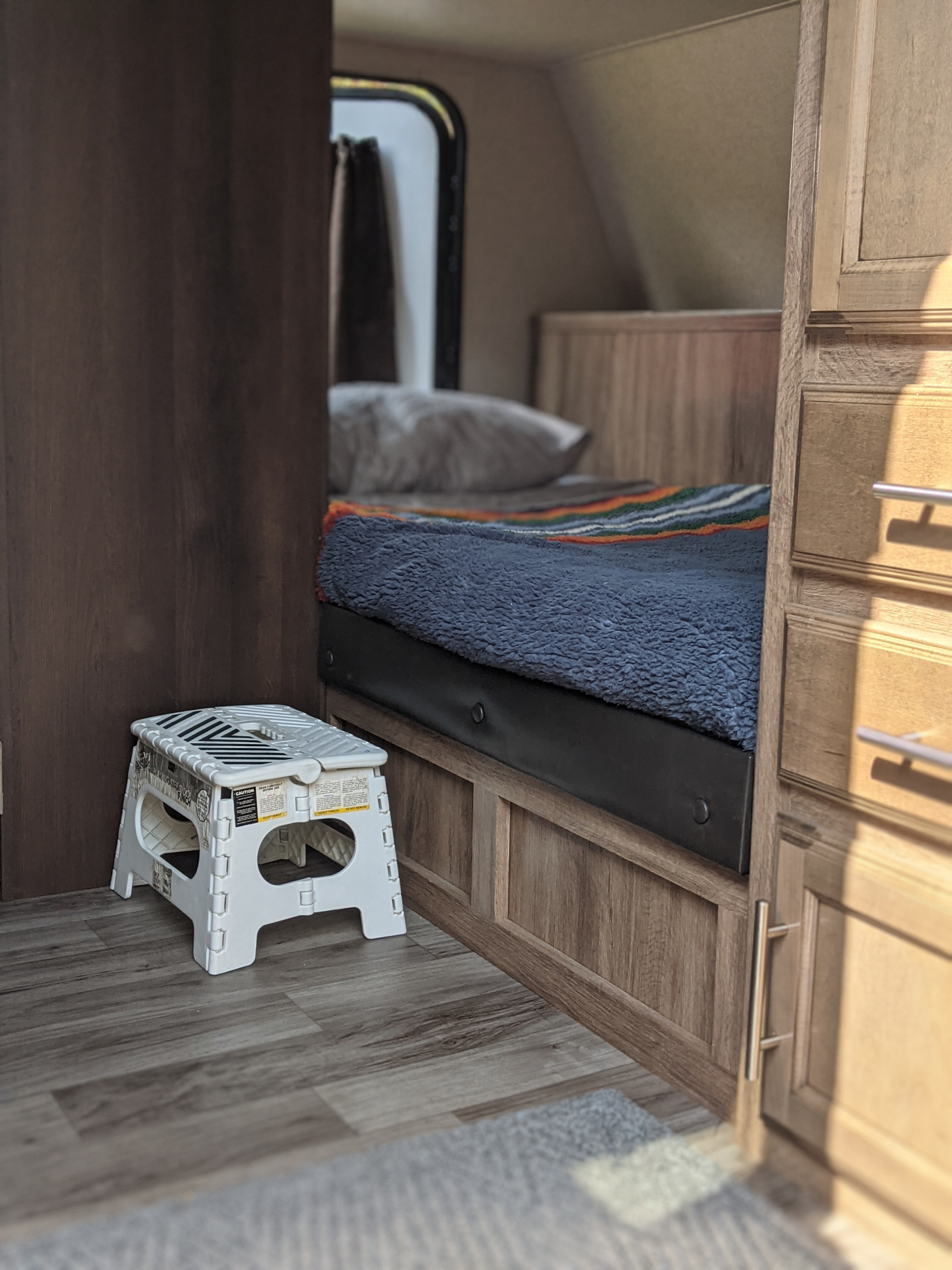 This cozy RV rental interior features a bed with a colorful blanket, a helpful step stool, and warm wood accents.. Jayco Jay Feather x213 2019