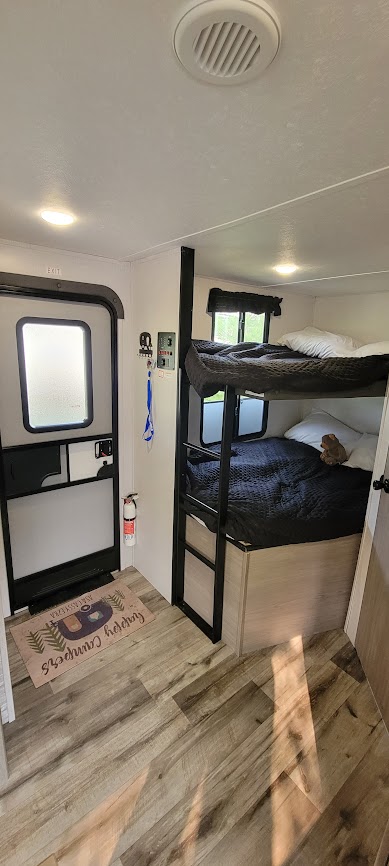 RV rental interior: inviting bunk beds, entry door with fire extinguisher, and a 'Happy Campers' mat on wood-look floor.. Dutchmen Coleman Light 2022