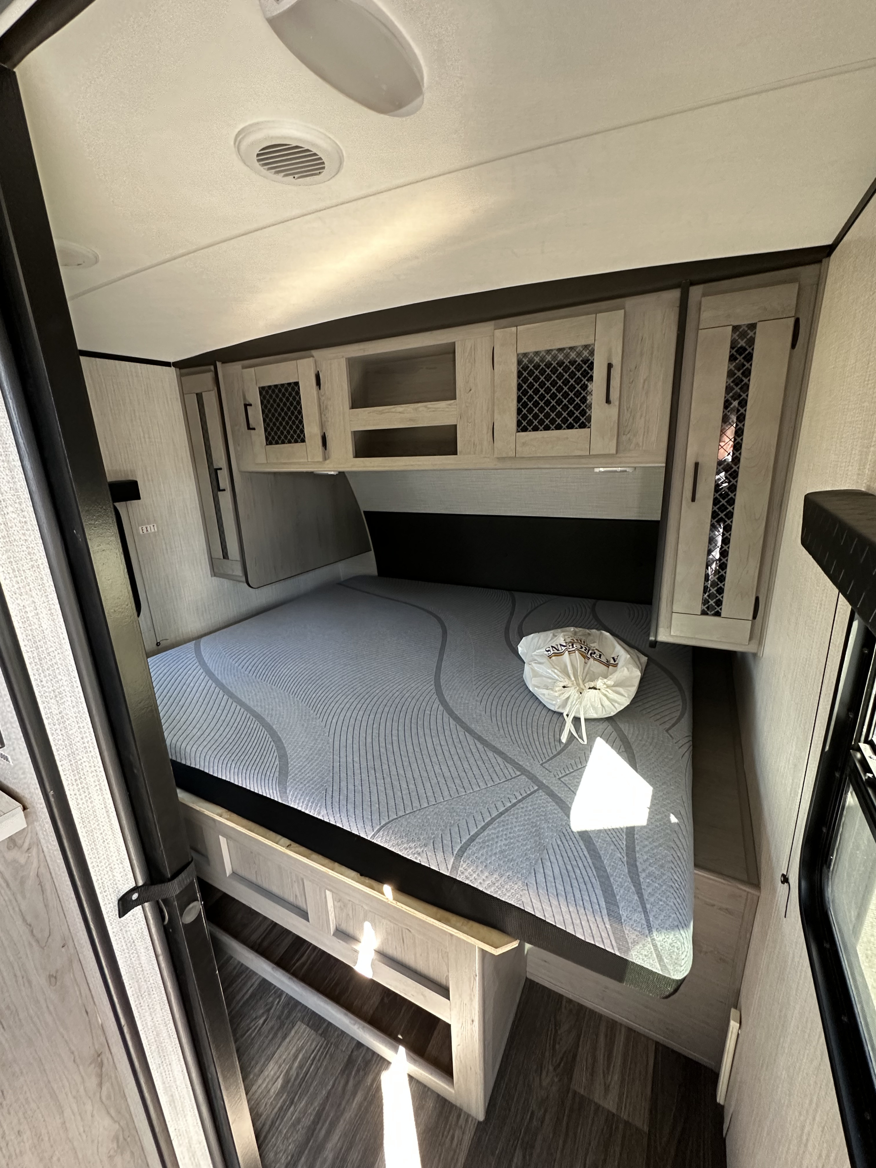 Inside this RV rental: a compact bedroom featuring a grey patterned mattress, overhead wood cabinets, and an EXIT sign.. Heartland RVs Mallard 2022