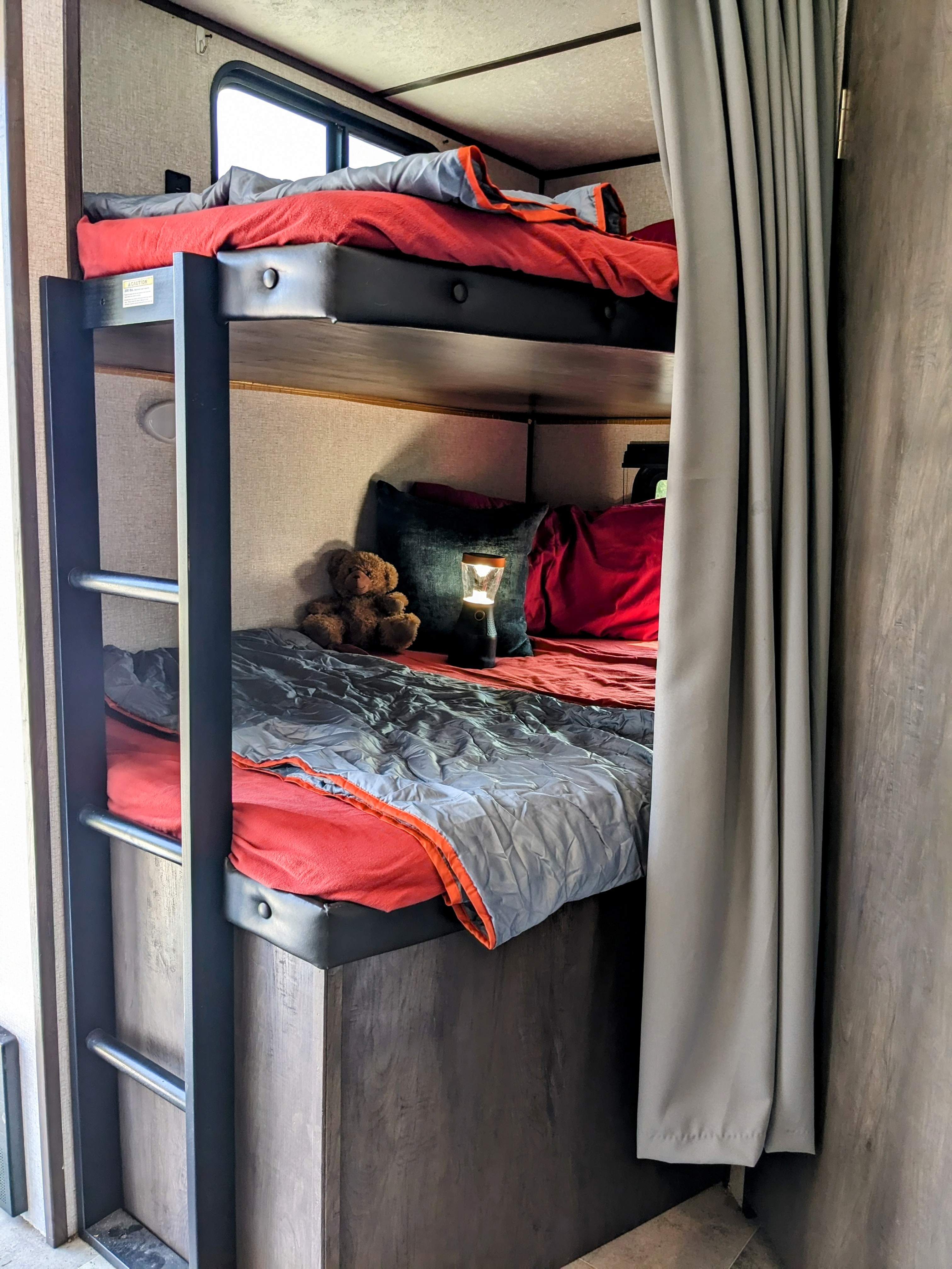 Cozy RV rental bunk beds with red sheets, gray comforters. A teddy bear and lantern sit on the lower bunk.. Coleman Lantern 2021