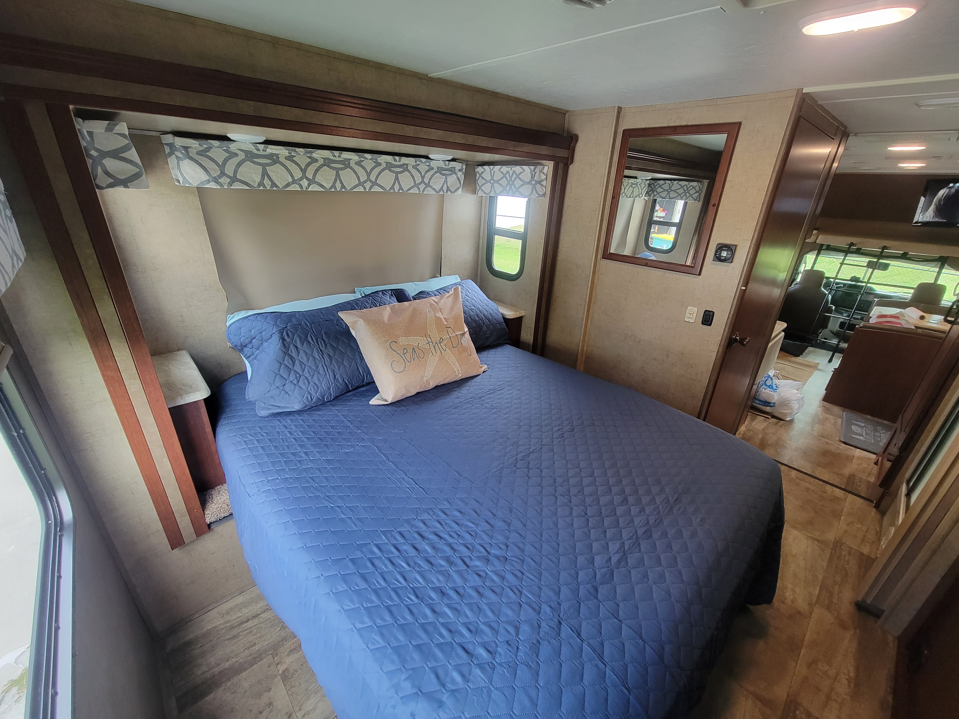 Inside a modern RV rental: a neat bedroom with blue bedding, windows, and mirror. Ahead is the driver's cabin.. Forest River Sunseeker 2017