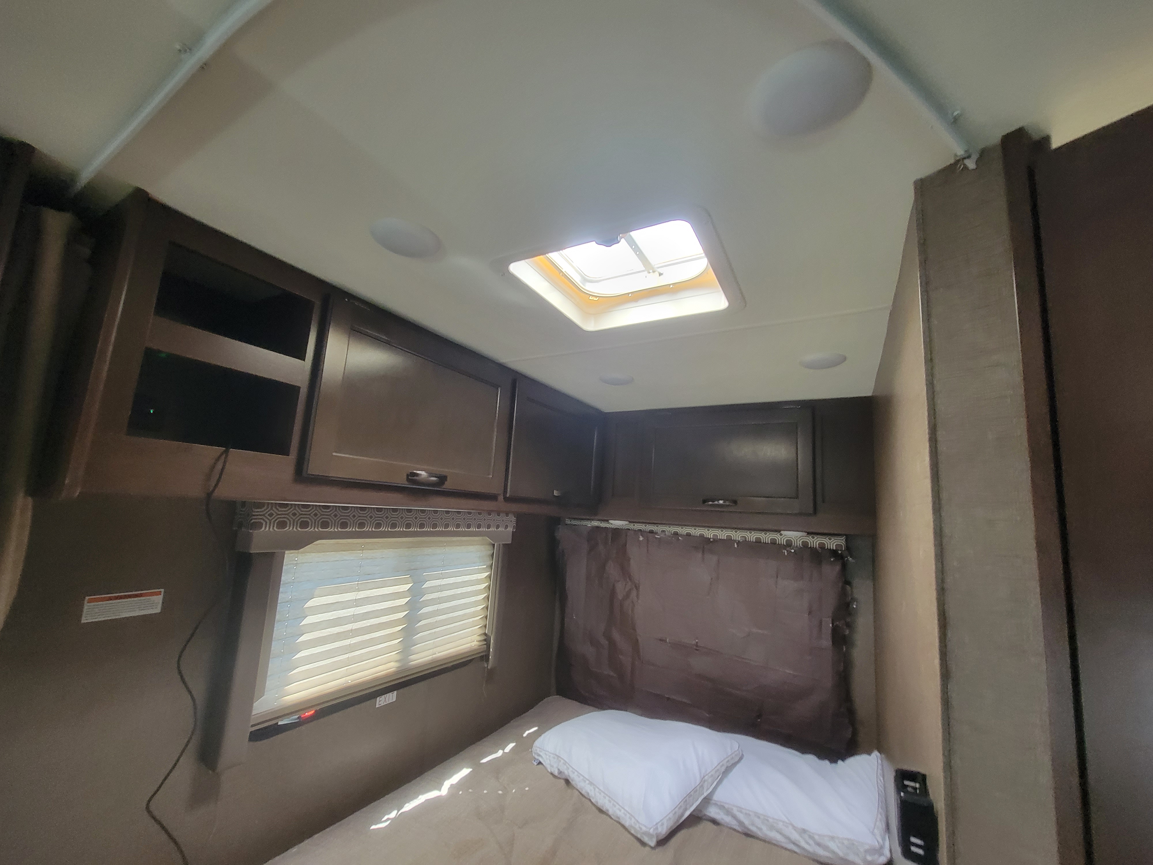 This cozy RV rental bedroom showcases a compact living space with a bed, overhead cabinets, window with blinds, and ceiling skylight.. Thor Four Winds 2020