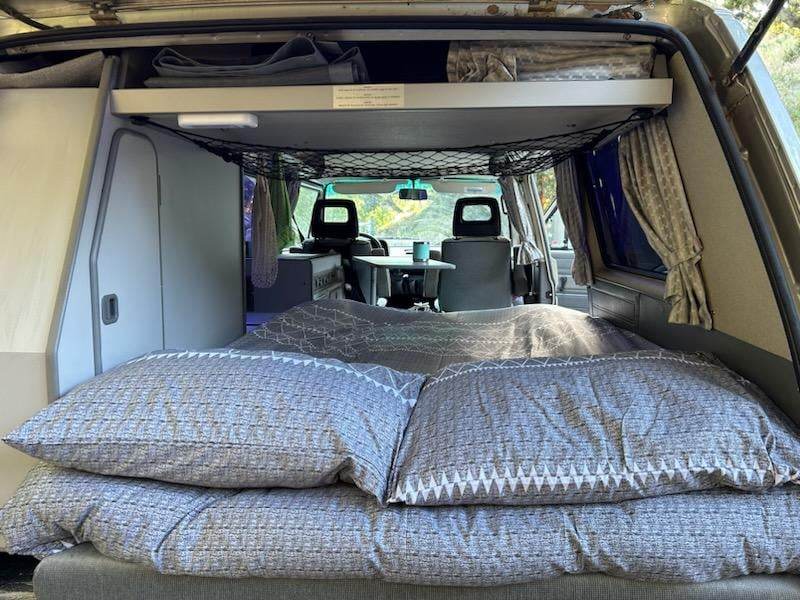 An inviting RV rental interior: a cozy bed with pillows, dining area, and storage for comfortable road trips.. Volkswagen T3 Westfalia 1990
