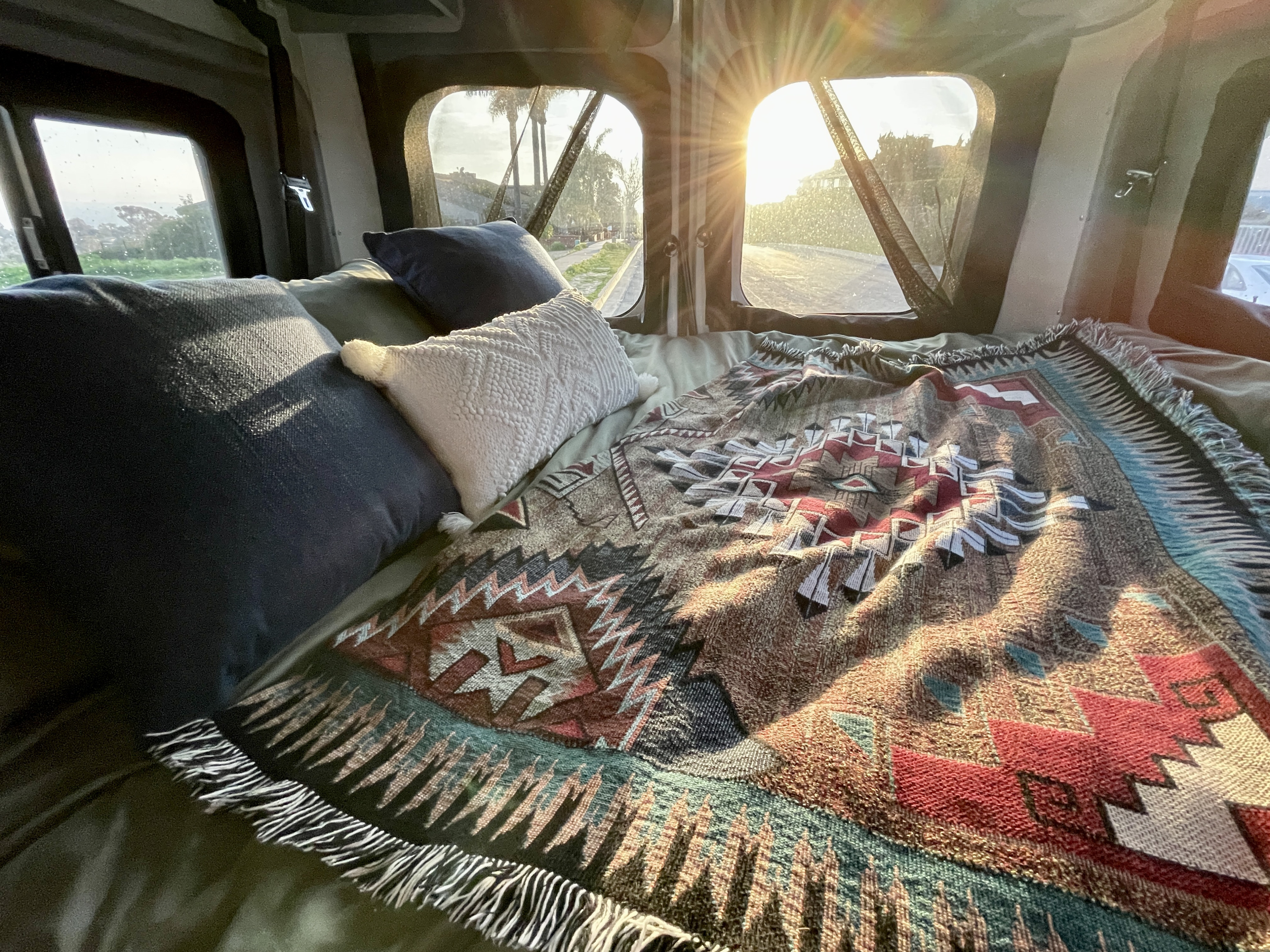 Cozy RV rental bed with pillows & patterned blanket, bright sun shining through windows, offering a relaxing road view outside.. Winnebago Solis 2021
