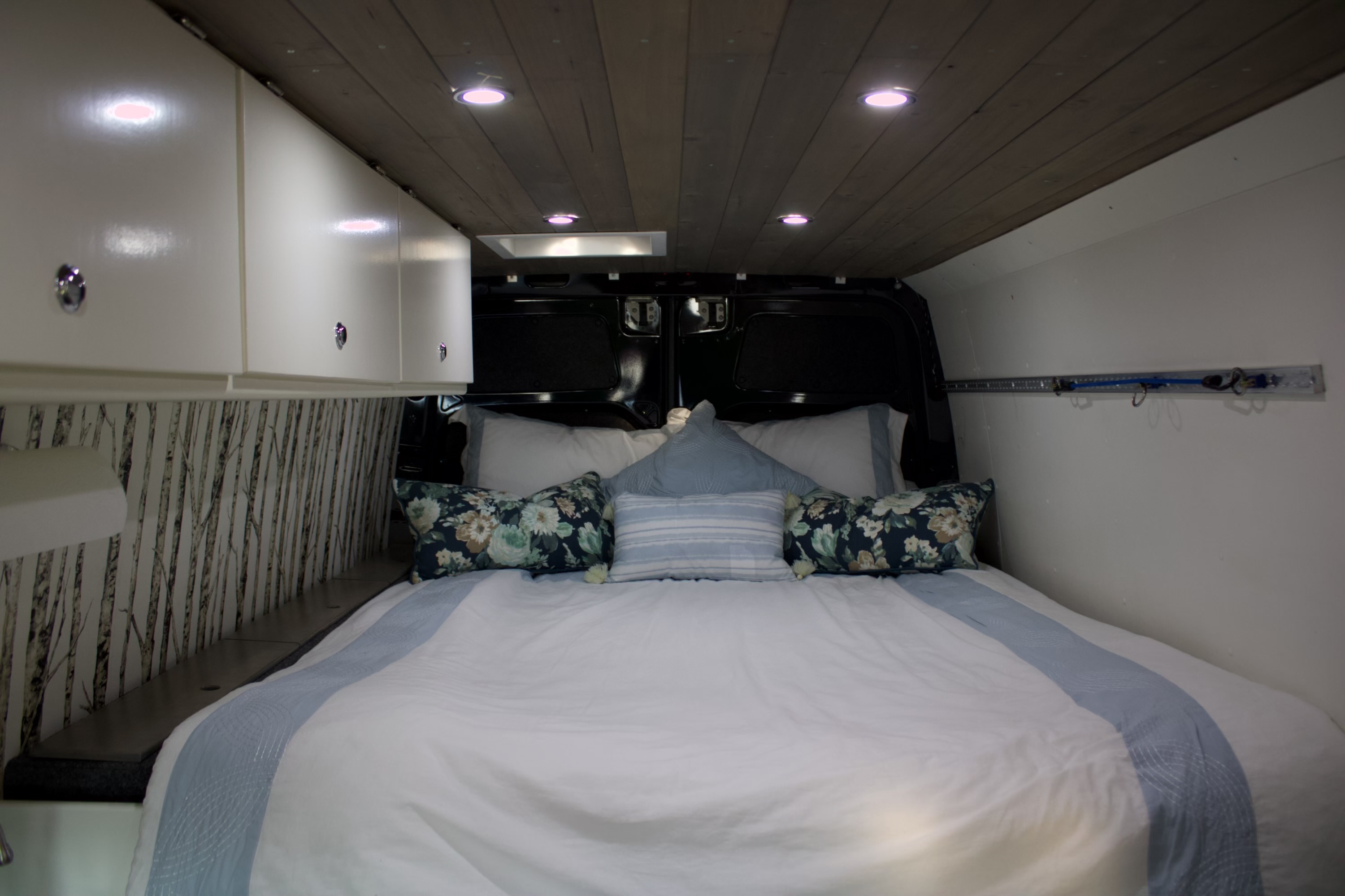 Cozy RV rental bedroom with a comfy bed, white bedding, floral pillows, wood ceiling, and birch tree wall decor.. Mercedes Sprinter 2012