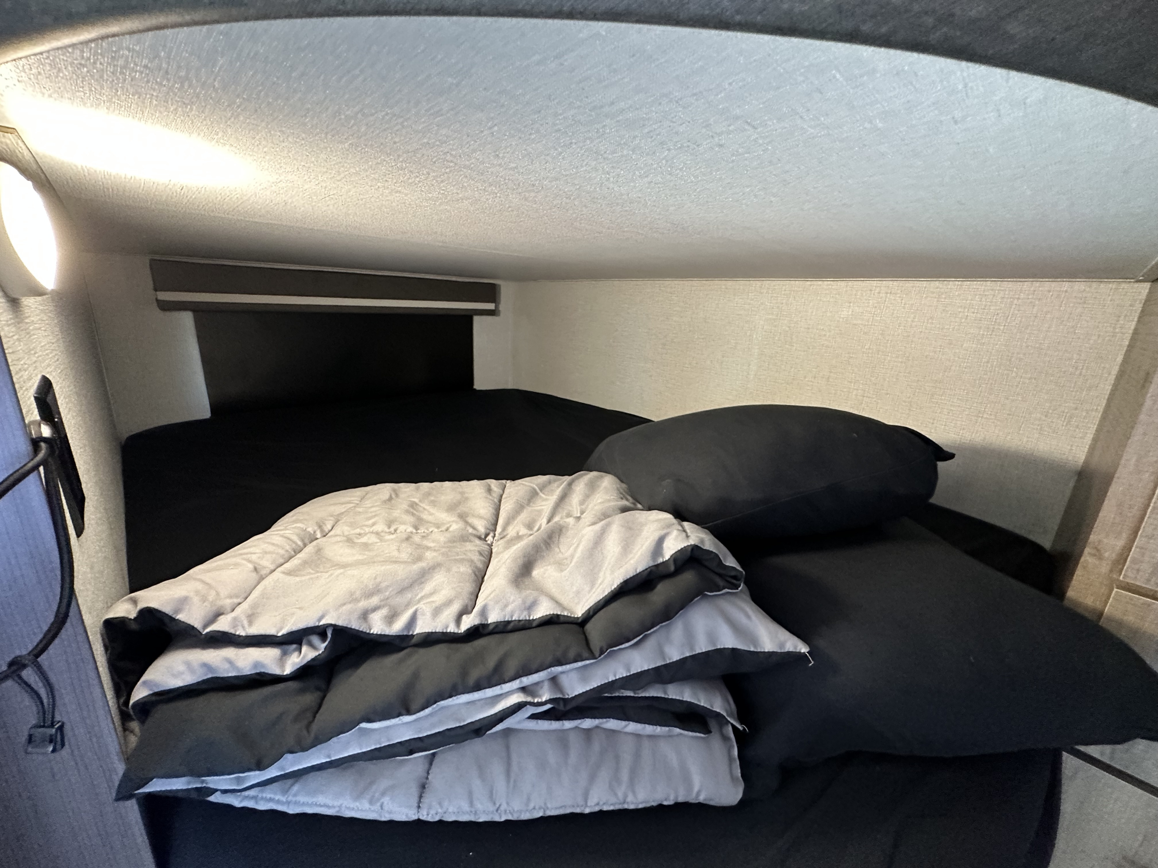 Cozy RV rental bunk bed with black sheets, pillow, and folded grey comforter, illuminated by an overhead light.. Grand Design Imagine 2023