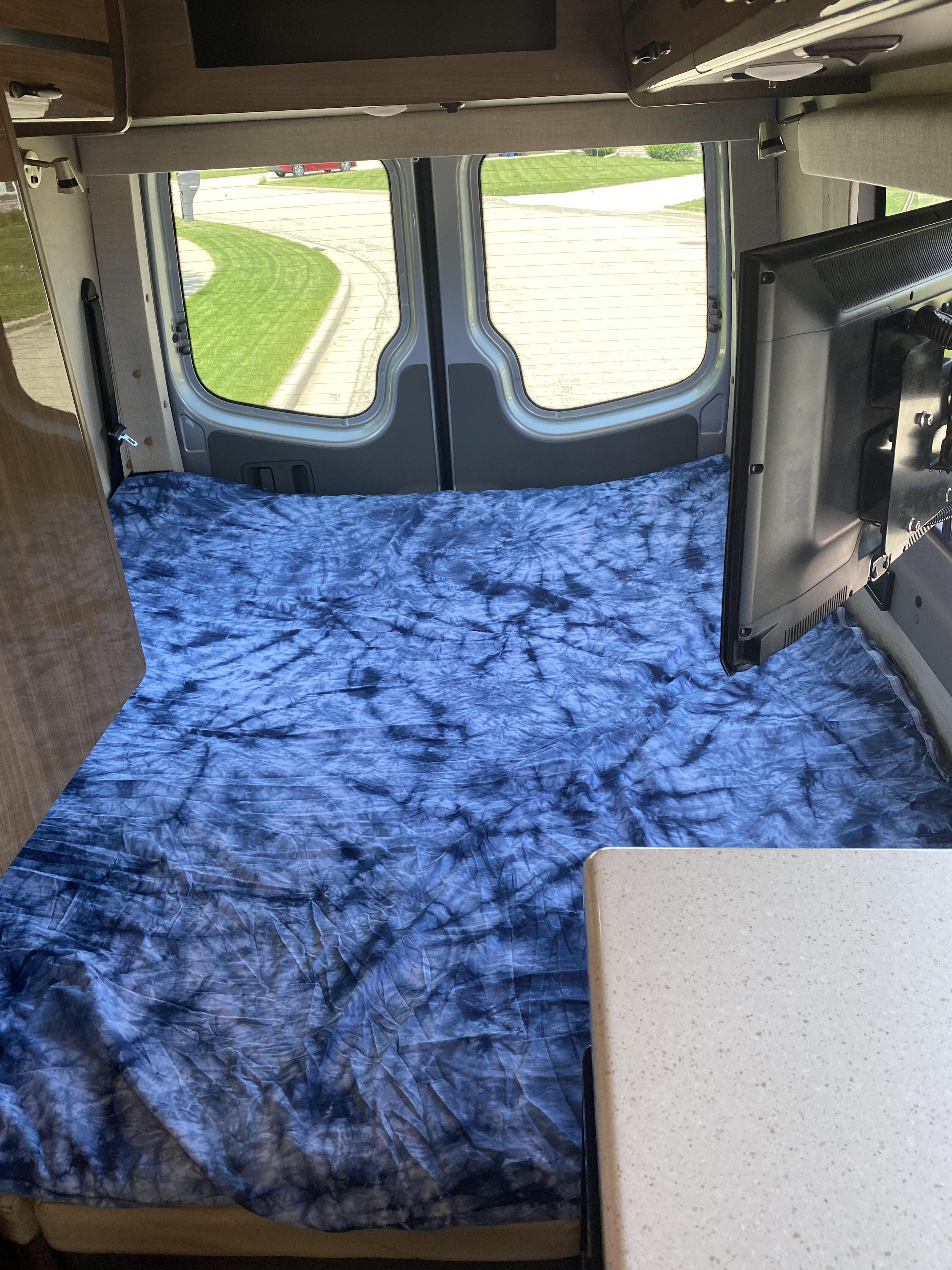 Inside an RV rental: a blue tie-dye bed fills the back, with a TV, kitchen counter, and rear windows showing a road.. Winnebago ERA 170X 2014