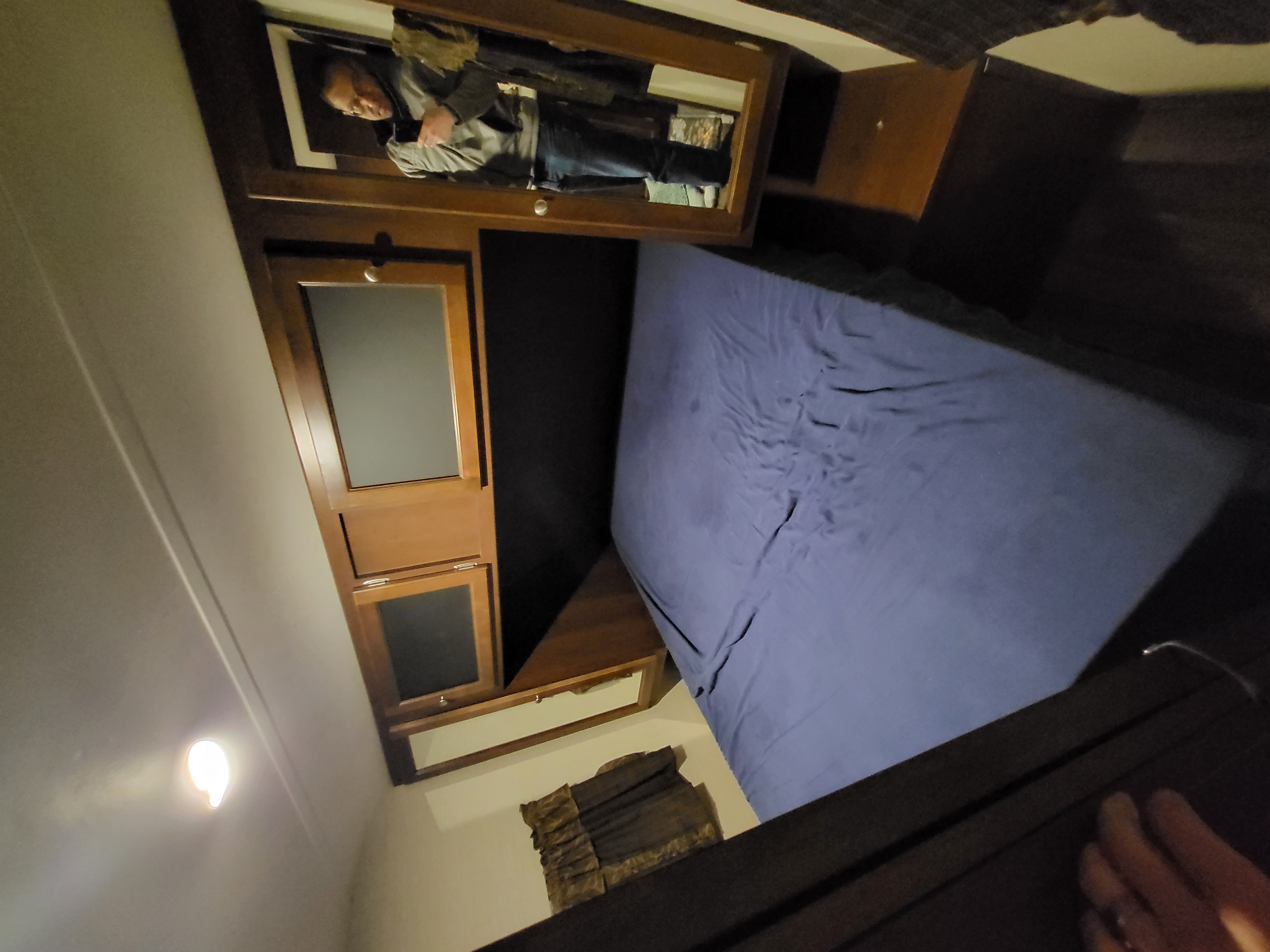 RV rental interior: A compact bedroom with a blue-sheeted bed, wood cabinets, and a person reflected in a wall mirror.. Keystone RV Bullet Ultra Lite 2016