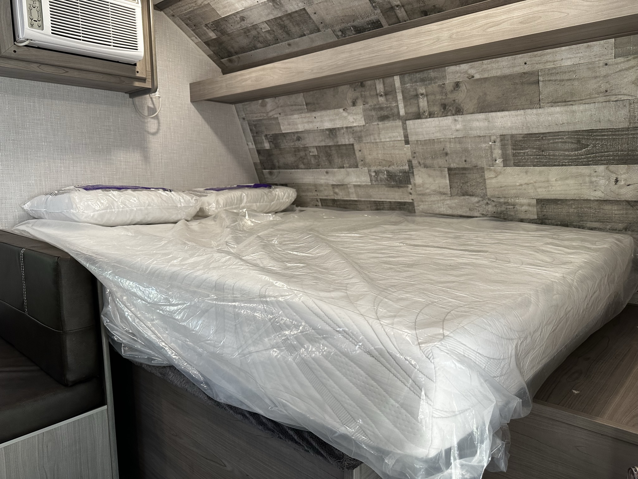 Bright RV rental interior showing a new bed with mattress in plastic, pillows, and AC unit. Modern wood paneling.. Dutchmen Coleman Lantern LT 2023