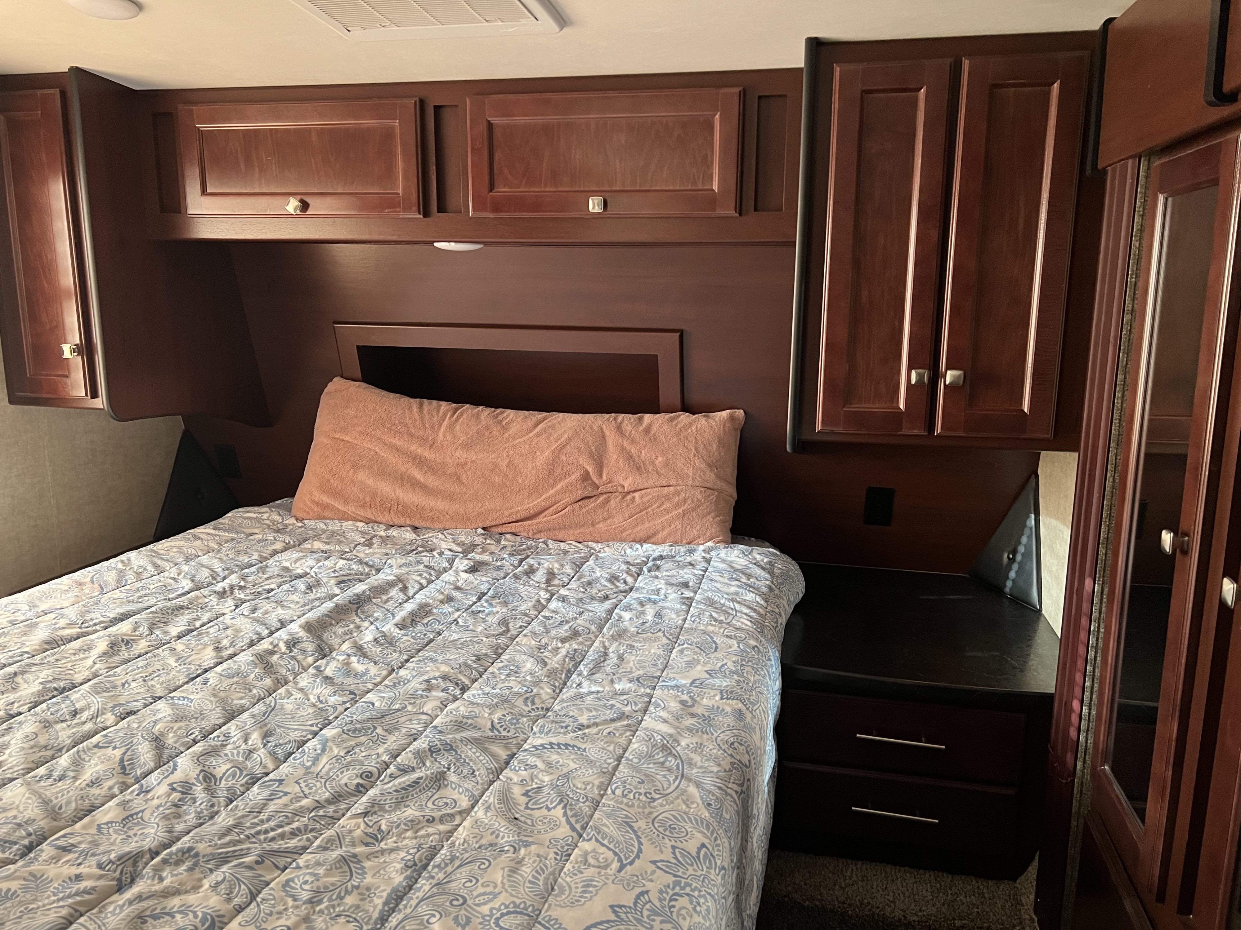 This RV rental bedroom features a comfortable bed with a patterned comforter, dark wood cabinets, and built-in storage.. Heartland RVs Road Warrior Toy Hauler 2015