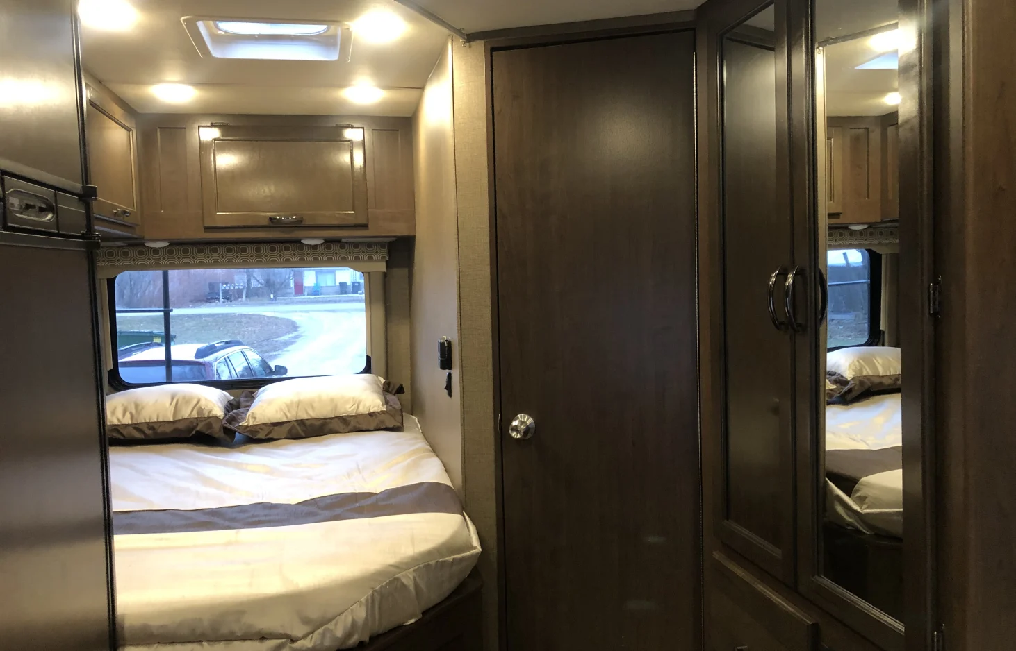 RV rental's inviting bedroom: a comfortable bed with pillows, overhead cabinets, window, and mirrored wardrobe.. Thor Four Winds 2018