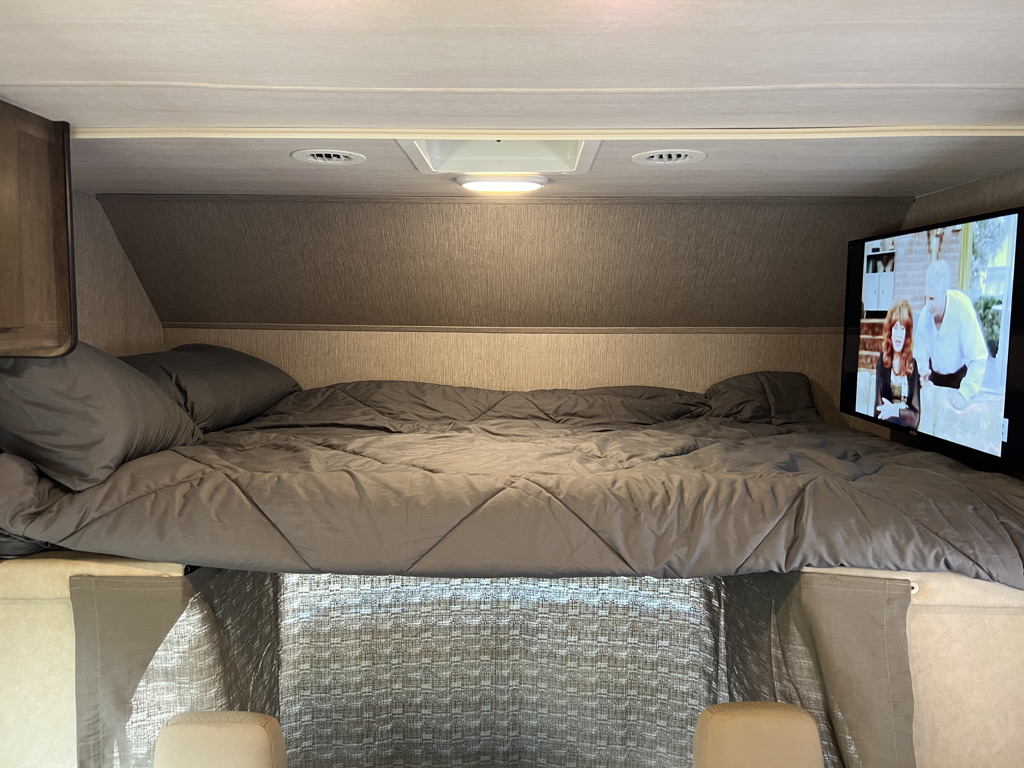 Inside an RV rental: a snug sleeping alcove with a large bed, grey duvet, pillows, and a wall-mounted TV.. Nexus Triumph 23T 2020