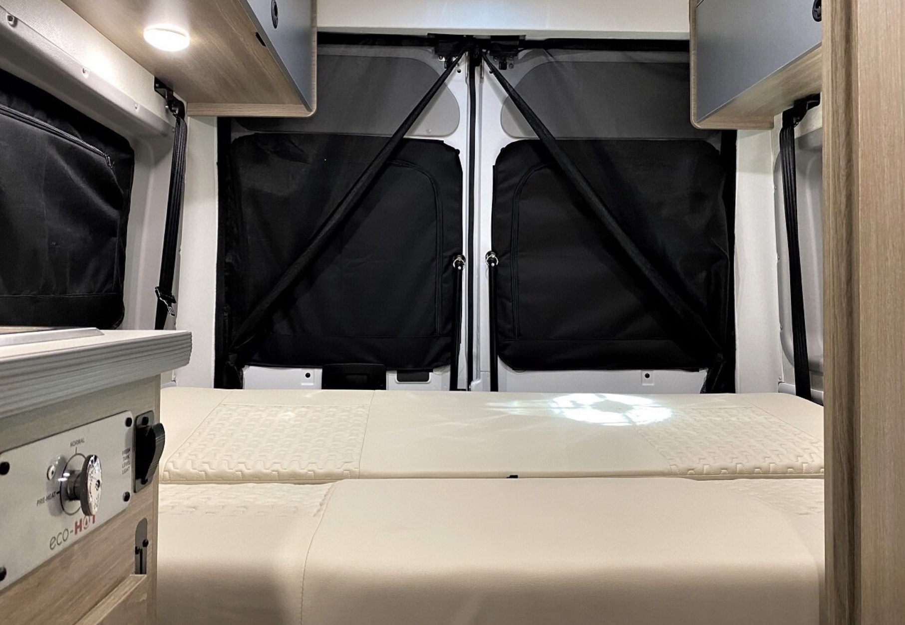 RV rental interior: A cozy bed faces rear doors with black window covers. A control panel, "eco-Hot," is on the left.. Winnebago Solis 2022