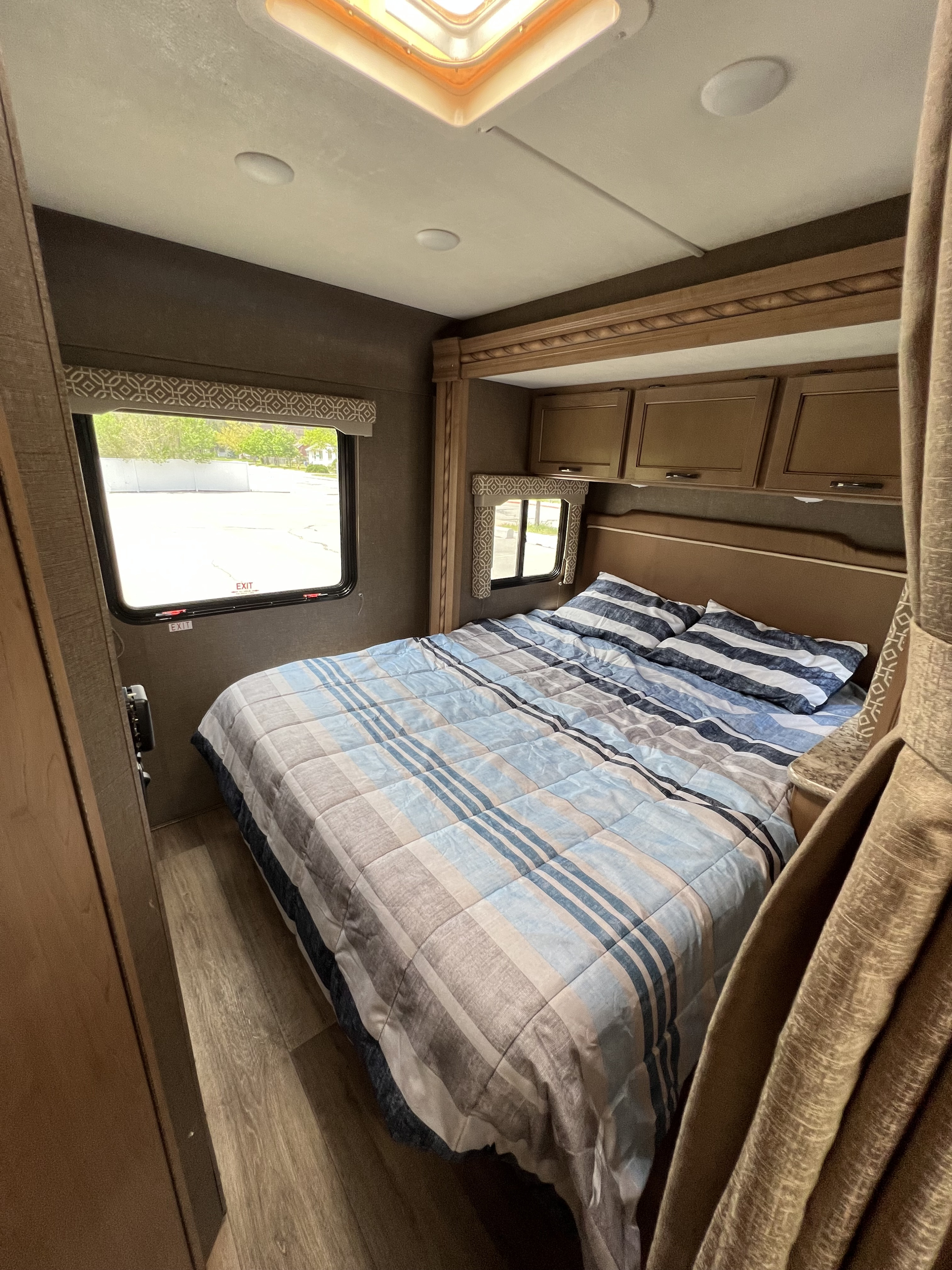 Inside a comfortable RV rental bedroom, showing a striped bed, side windows, and overhead storage cabinets.. Thor Motor Coach Freedom Elite 2018