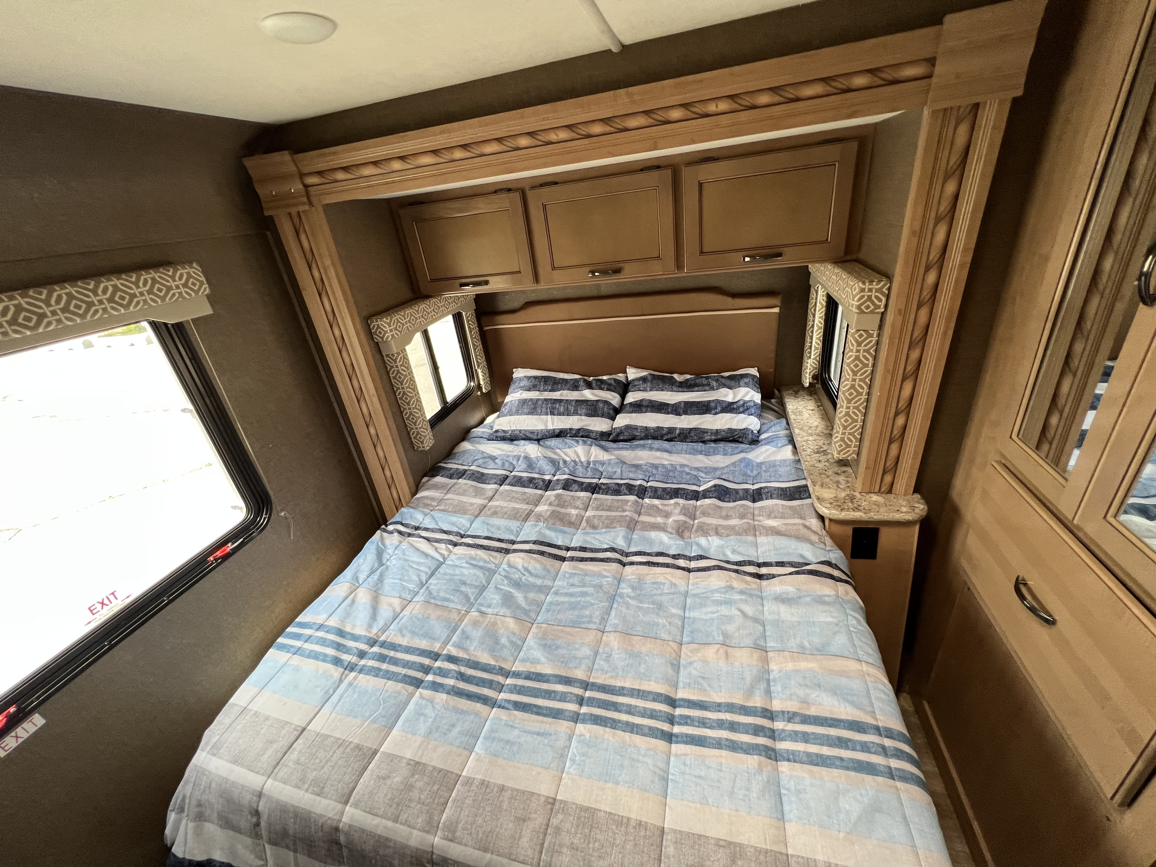 Interior RV rental bedroom featuring a bed with blue and gray striped bedding, wooden overhead cabinets, and a side window.. Thor Motor Coach Freedom Elite 2018