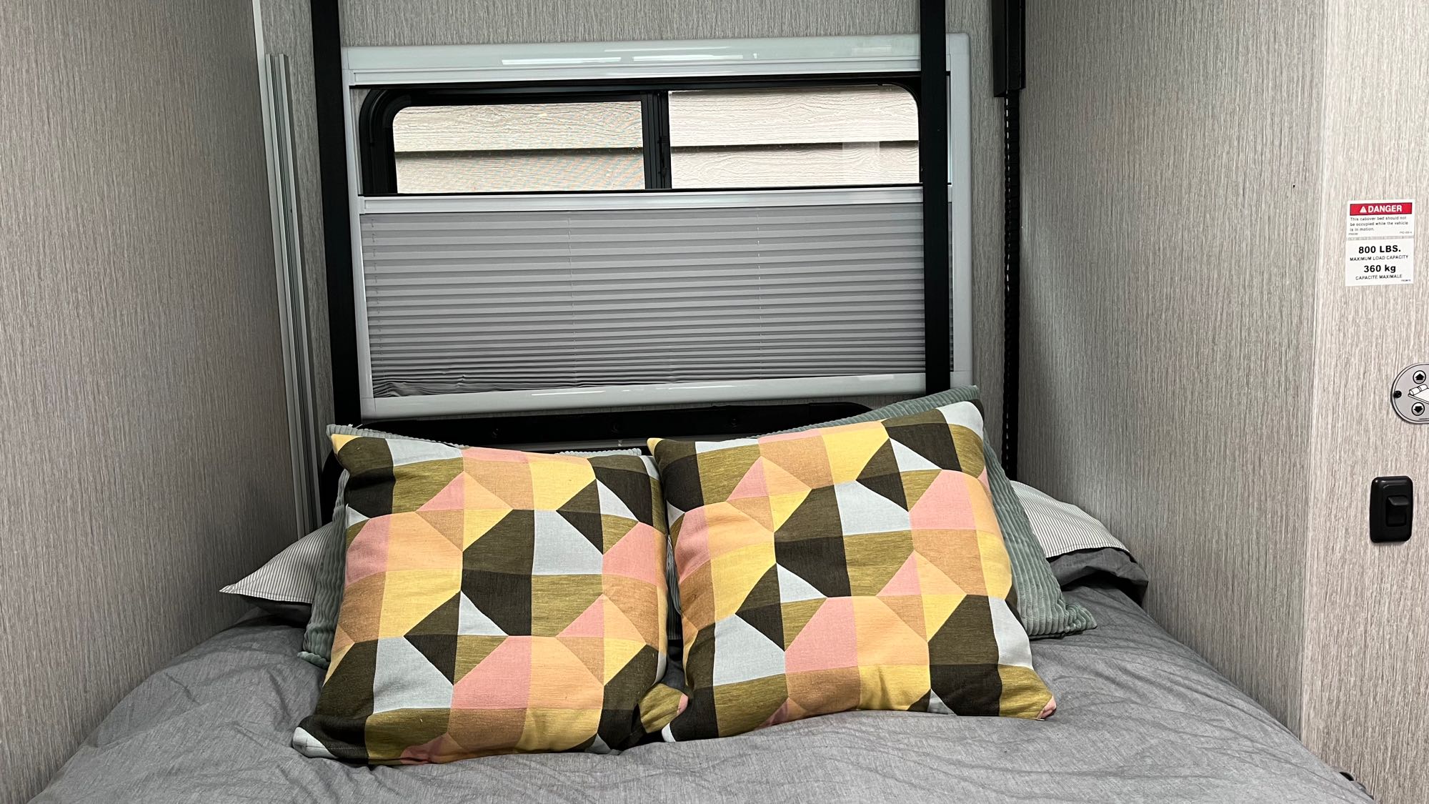 Cozy RV rental bed with colorful geometric pillows, window shades, and a rollover bed safety warning.. Forest River Coachmen Cross Trail XL 2023