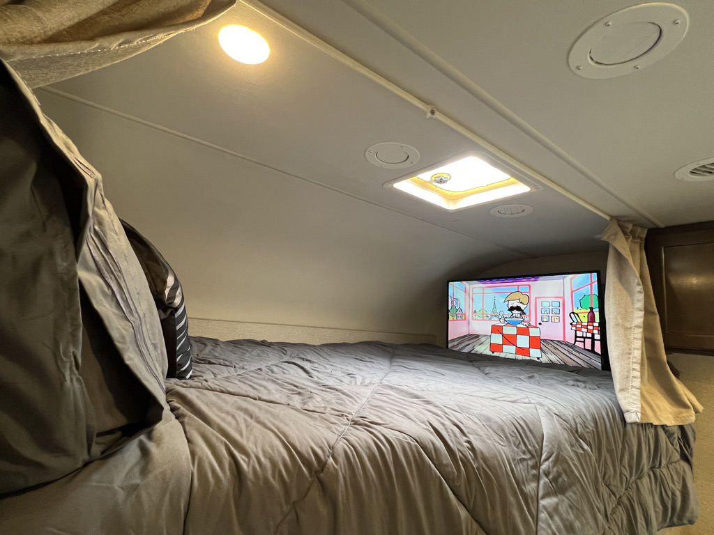 Inside a cozy RV rental bunk: a large bed with gray comforter, TV playing animation, and overhead lights.. Thor Motor Coach Freedom Elite 2017