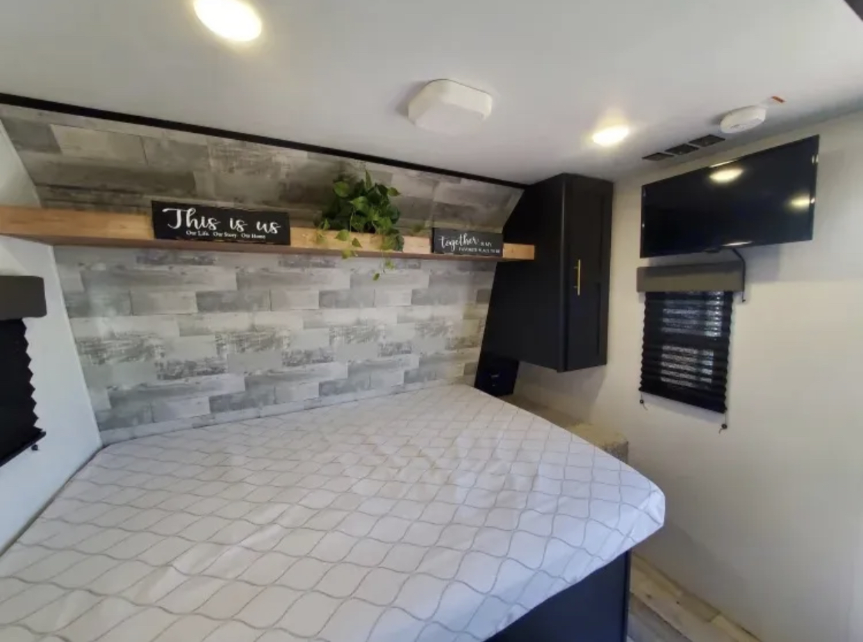 Cozy RV rental bedroom, featuring a patterned bed, decorative wood-paneled wall, storage, TV, and window for comfortable travel.. Keystone RV Springdale 2022