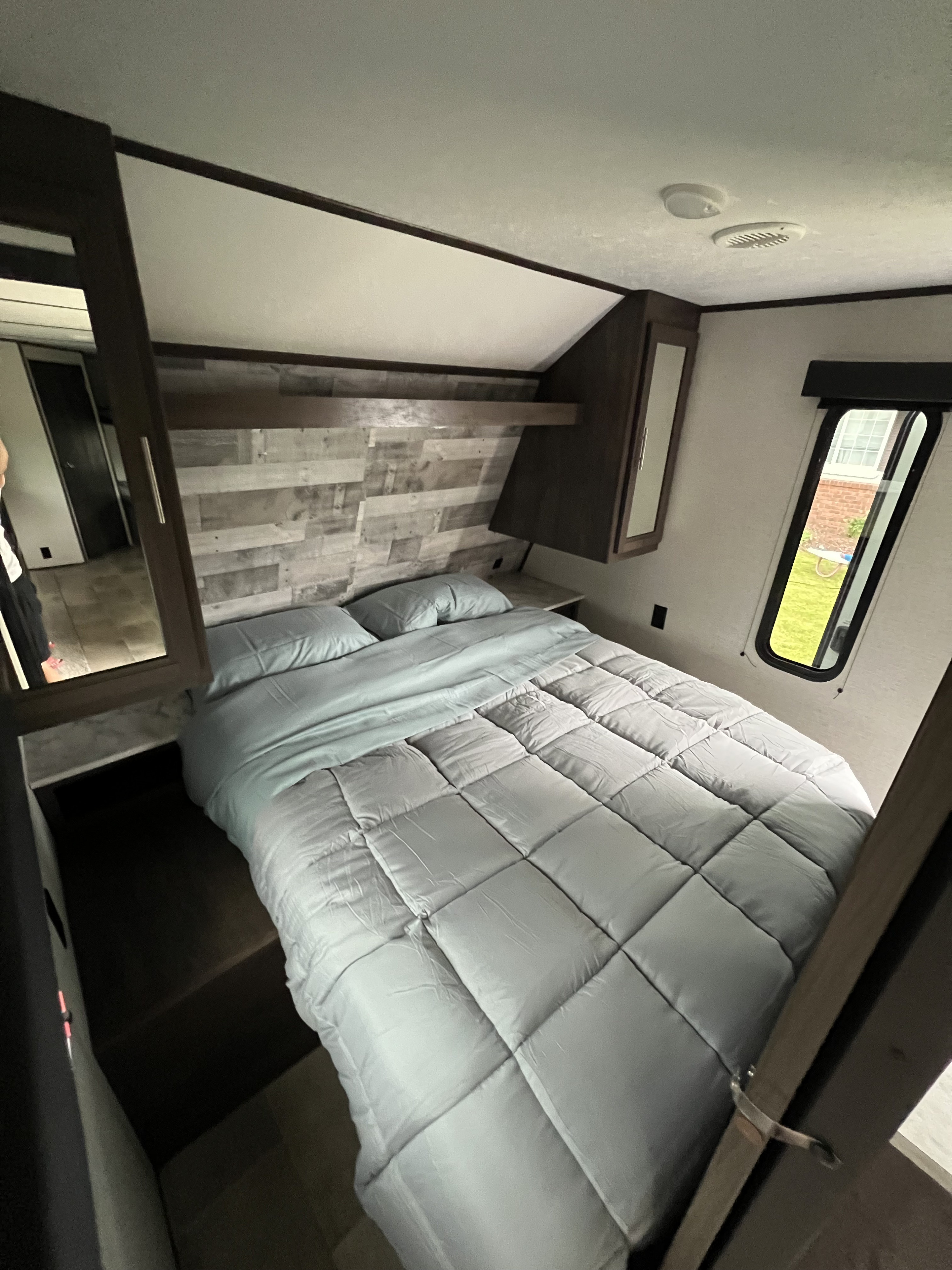 Relaxing RV rental bedroom with a comfy light blue bed, wood-paneled wall, and a side window, providing a compact, cozy space.. Coleman dutchmen 2021