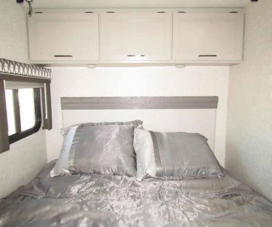 Cozy RV rental bedroom featuring a comfortable bed with silver bedding, overhead cabinets, and a window for natural light.. Thor Quantum 2023