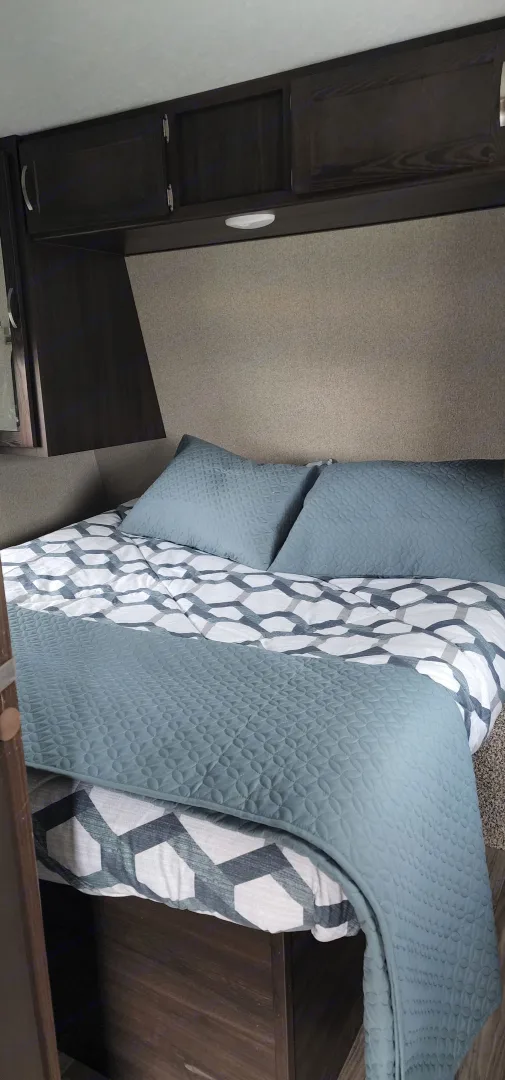 Cozy RV rental bedroom showcasing a bed with patterned duvet, soft teal blanket & pillows, and dark overhead cabinets.. Keystone RV Springdale 2019